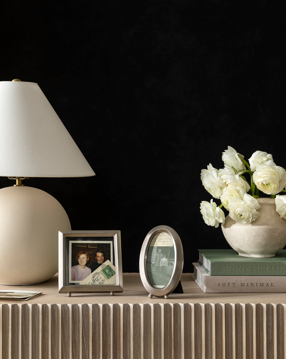 A beige table lamp, the Blanche Pewter Picture Frame, a vase of white tulips, and stacked books sit on a light wood console with iron corners against a black background.