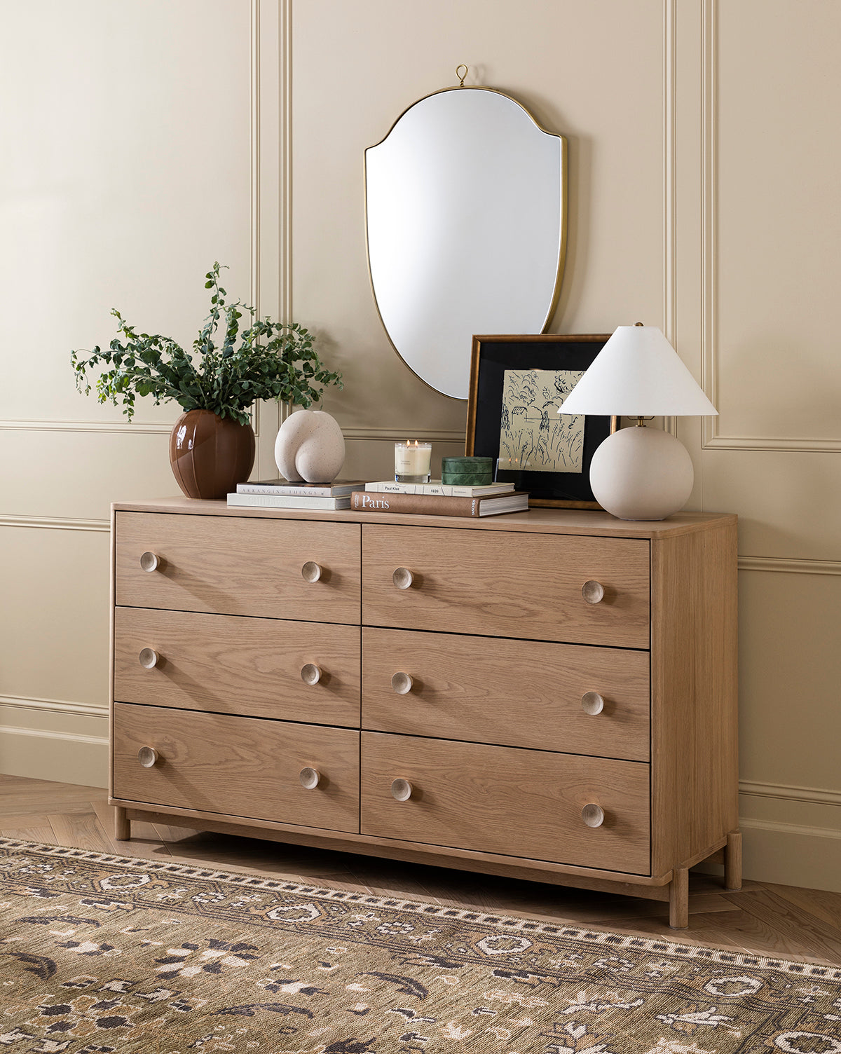 The Baxter Dresser, featuring six drawers, sits against a beige paneled wall with a round mirror above. Finished in oak, this transitional piece is styled with a lamp, books, greenery in a vase, and completes the bedroom with a patterned rug beneath, by McGee & Co.
