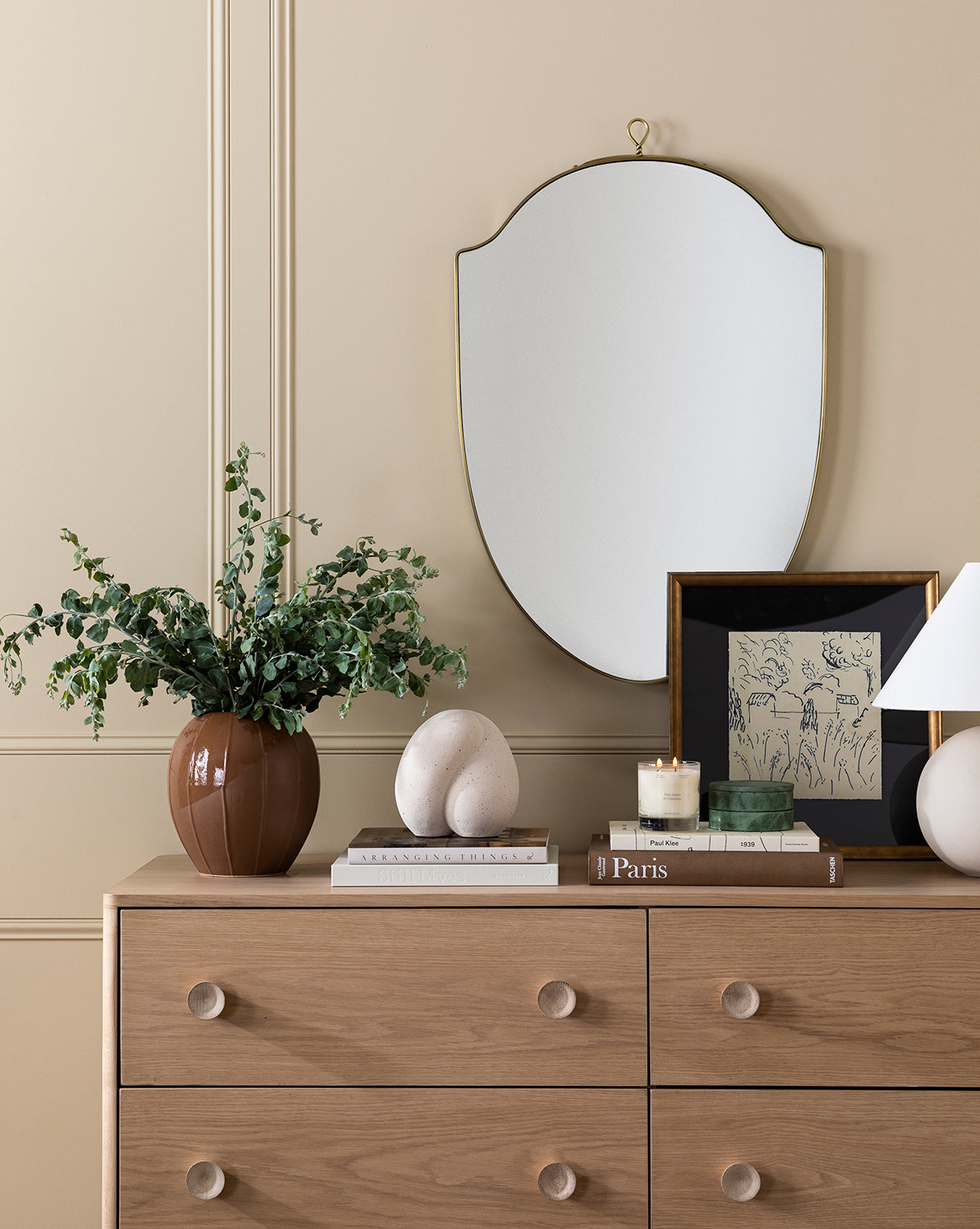 A wooden dresser displays a brown vase with greenery, sculpted spheres, books, a candle, framed art, and a small lamp. Above, the Allegheny Wall Mirror hangs on the beige paneled wall by McGee & Co.