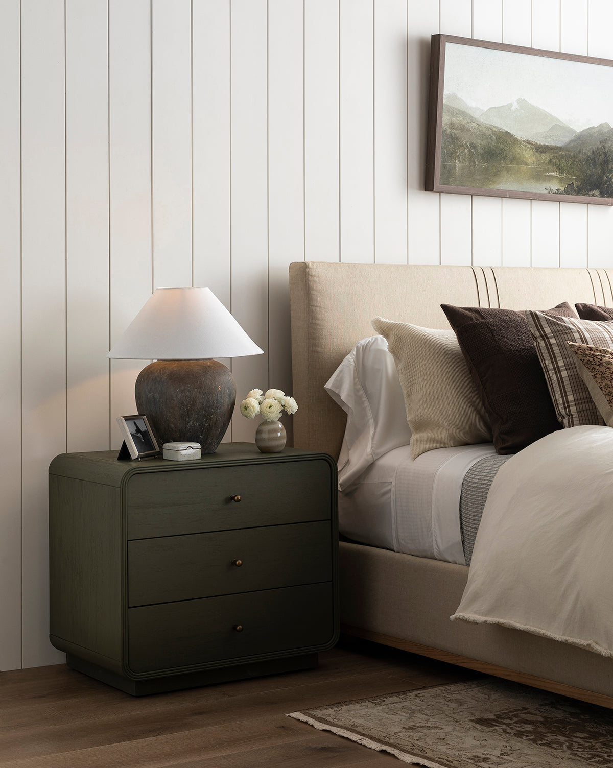 A cozy bedroom features a beige upholstered bed, layered pillows, and a green nightstand with a ceramic lamp, framed photo, and flowers. Above the bed on the shiplap wall hangs Darsey Lake Peaks in a walnut wood frame.