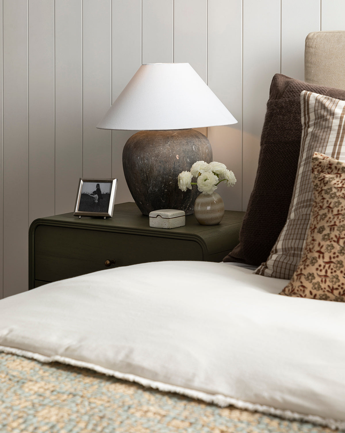 A cozy bedroom nightstand features a rustic lamp, the Blanche Pewter Picture Frame, a small vase of white flowers, a decorative box, and a bed with patterned and plain pillows set against a white paneled wall.