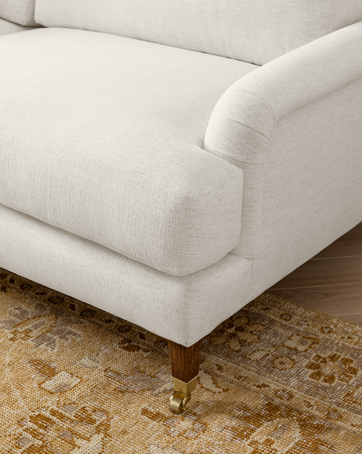 Close-up of the Lucille English Roll Arm Sofa 78 in cream, featuring down-wrapped cushions and wooden legs with brass caster wheels, set on a beige and light brown patterned rug by McGee & Co.