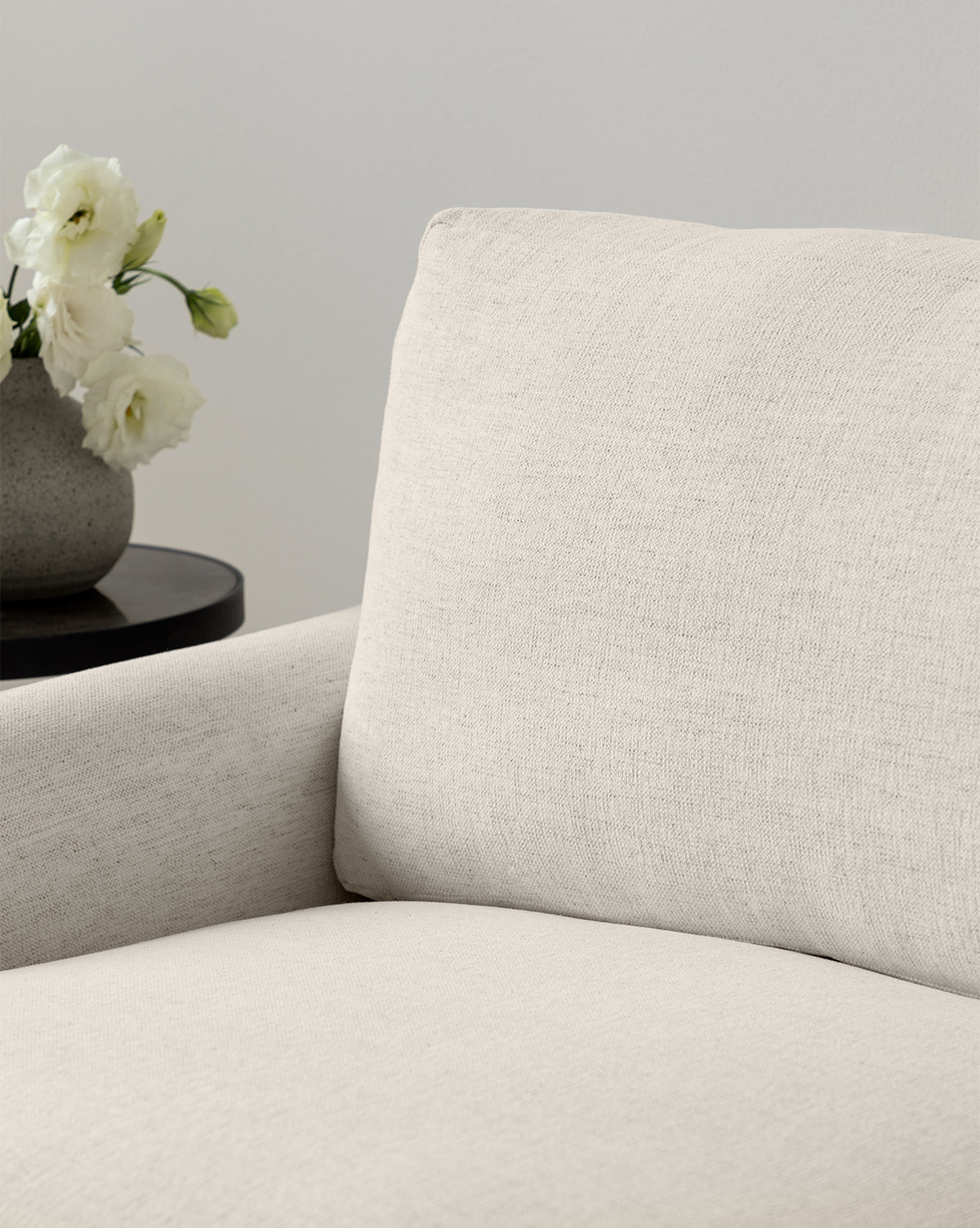 A close-up of the Lucille English Roll Arm Sofa 78 in light beige fabric with a textured surface and plush, down-wrapped cushions, by McGee & Co. In the background, a round dark side table holds a gray vase with white flowers.