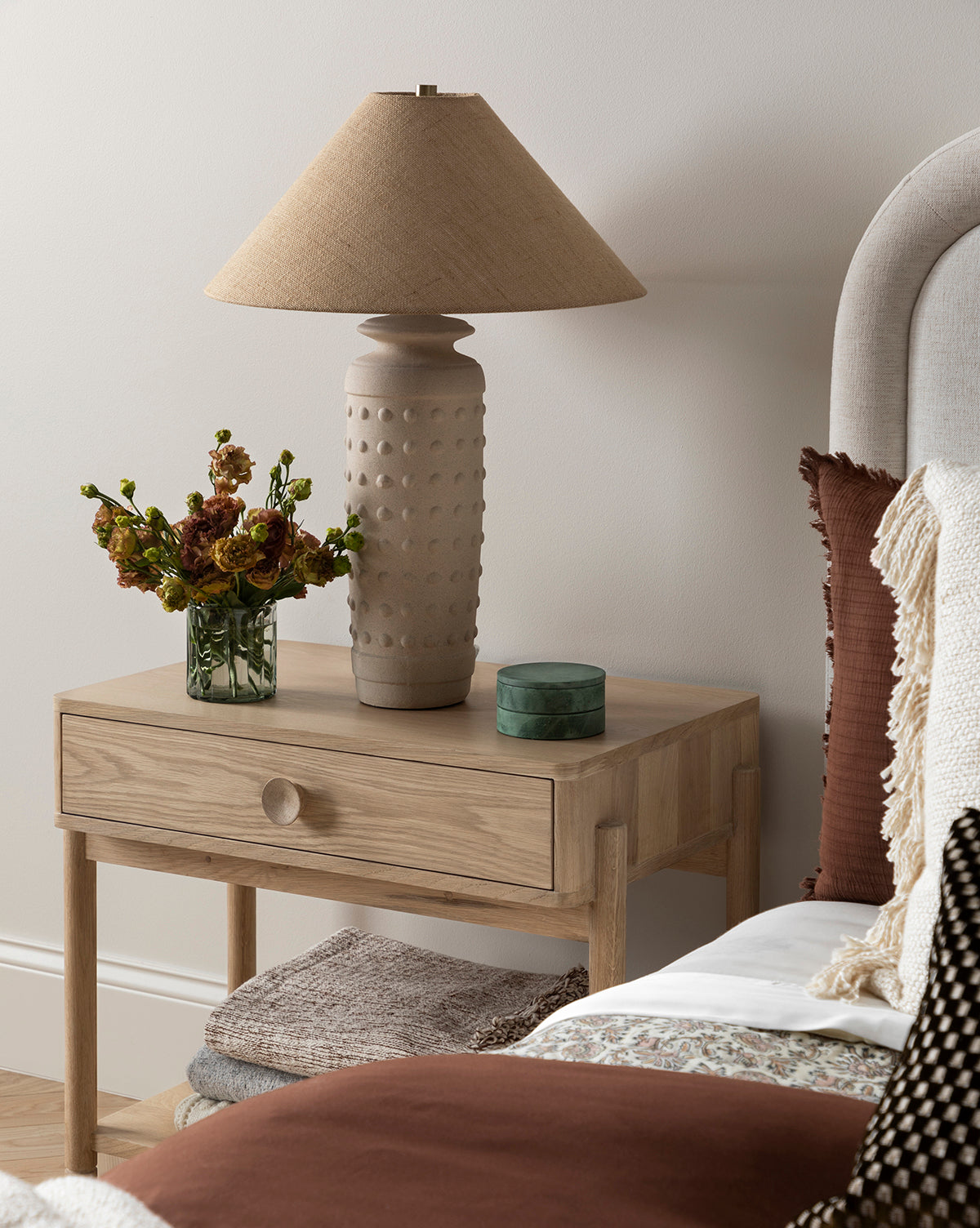 A wooden bedside table with the Sonoran Table Lamp, featuring a burlap shade, a small green box, orange flowers in a vase, and folded blankets below sits next to a bed with neutral and brown pillows and a cream throw blanket by McGee & Co.