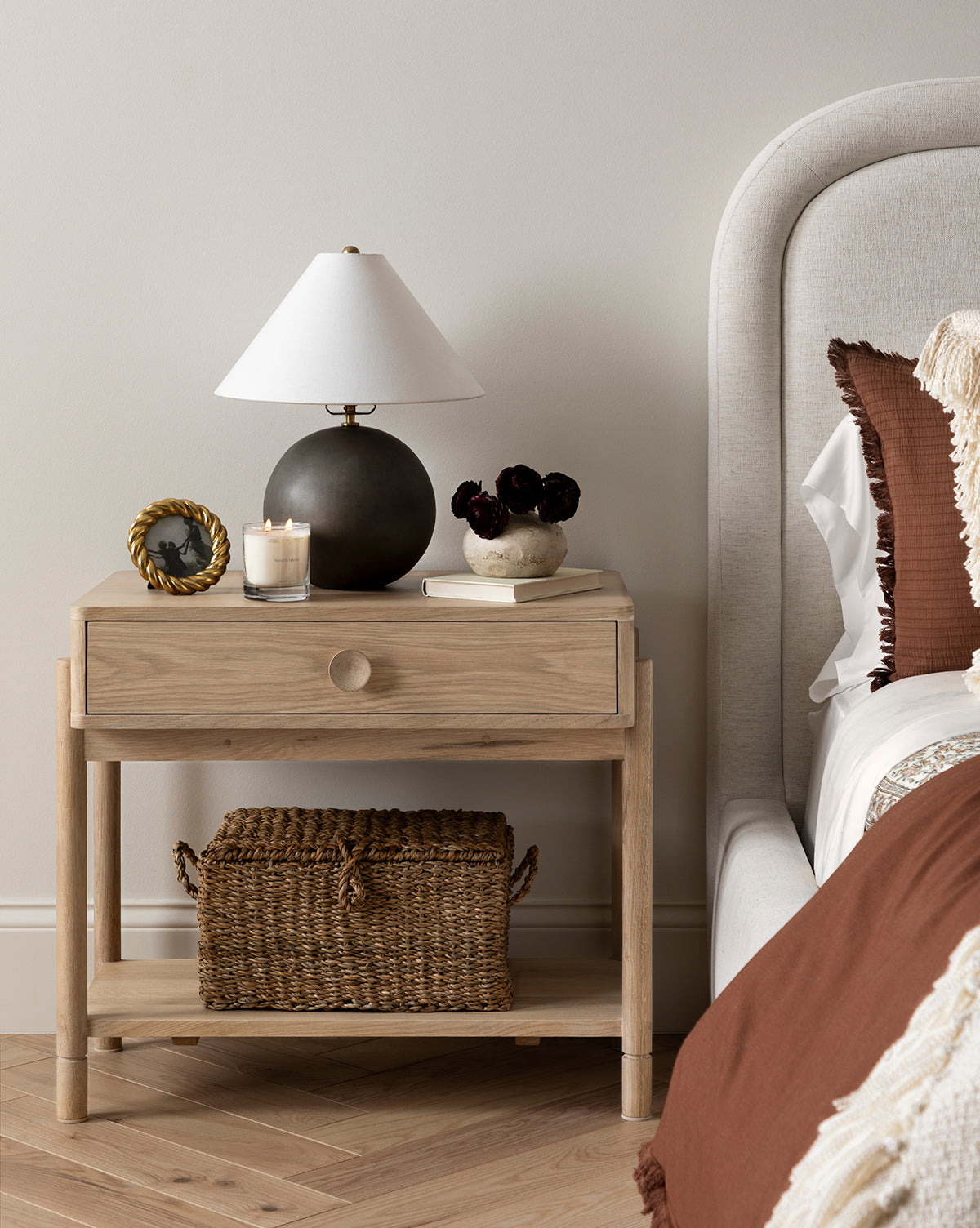 The Baxter Nightstand in light oak features a transitional design, topped with a round black lamp, picture frame, candle, and dark flowers. A woven basket rests on its lower shelf next to a beige upholstered bed with brown and cream pillows by McGee & Co.