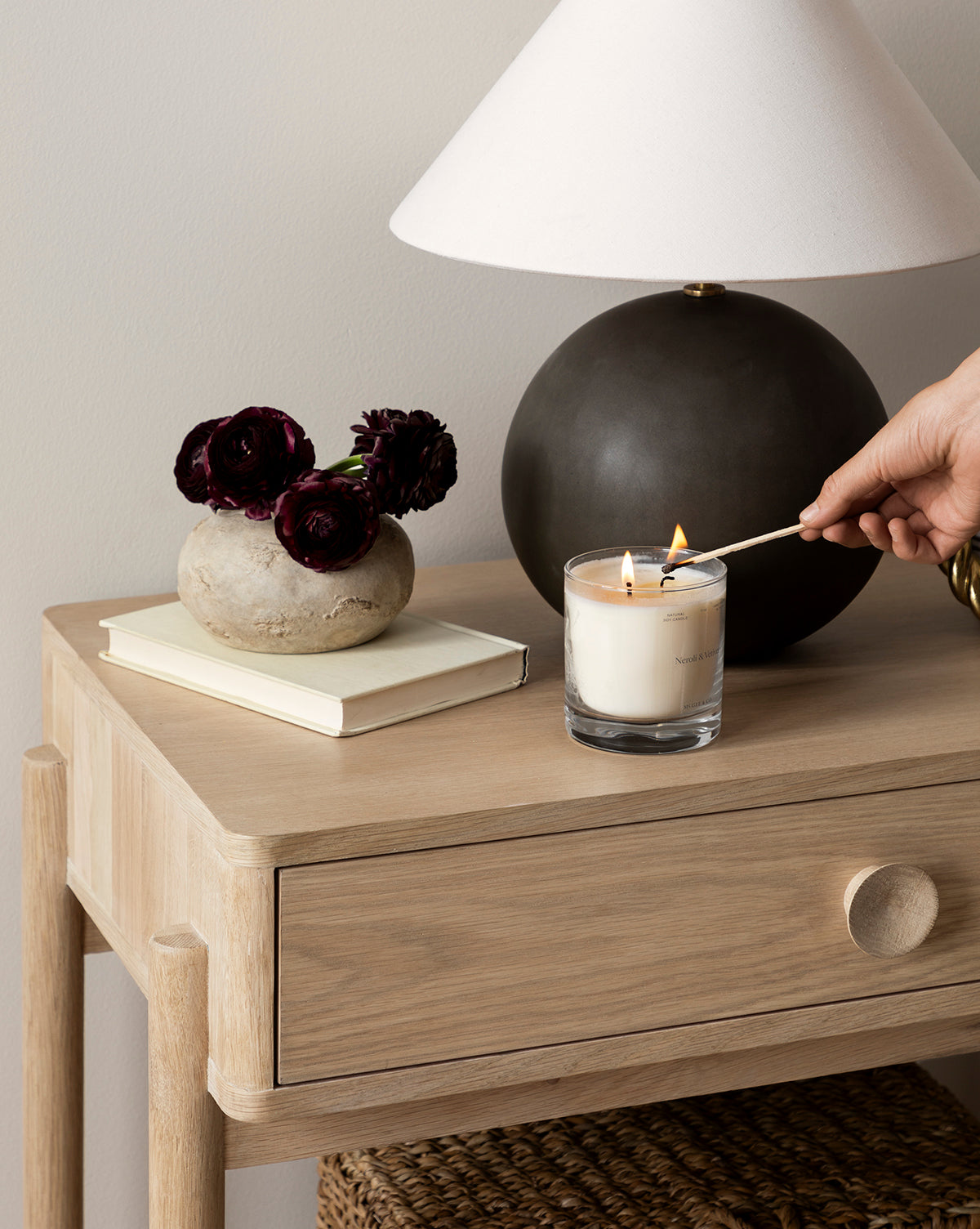 A hand lights a white candle in a glass jar on the Baxter Nightstand—perfect for transitional design—next to a closed book, a stone vase with dark flowers, and a black lamp with a white shade, by McGee & Co.