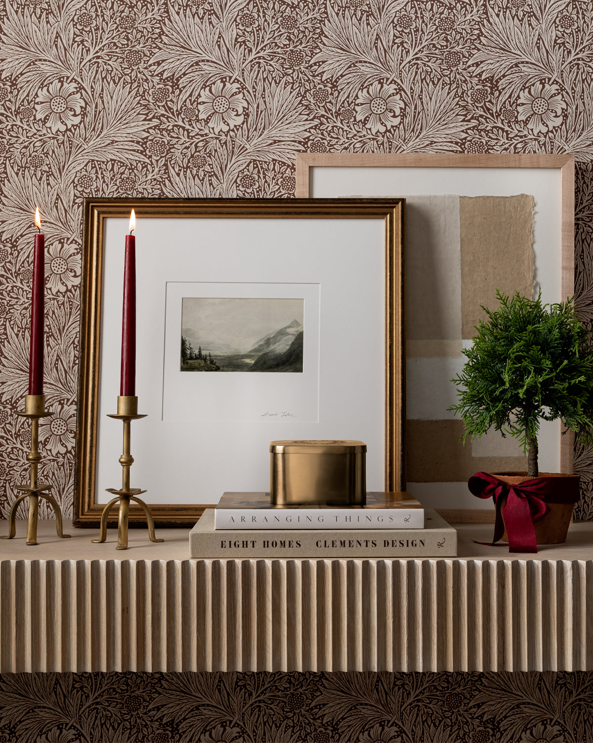A shelf displays two tall red candles in gold holders, stacked books, a gold tin, and a small potted plant with a red ribbon. The Window to the Mountains framed art stands out against patterned brown floral wallpaper.