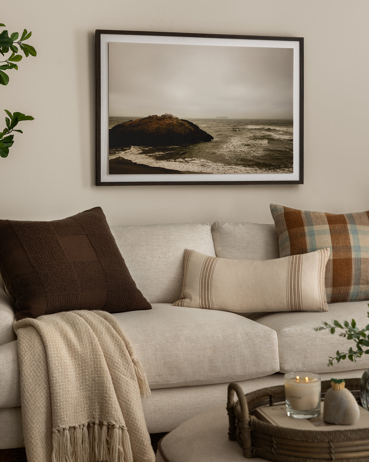 A cozy beige sofa with textured pillows and a knit blanket sits beneath the “Sunday Fog” photography giclee print in a walnut wood frame. On the coffee table in front, a tray holds candles and greenery.