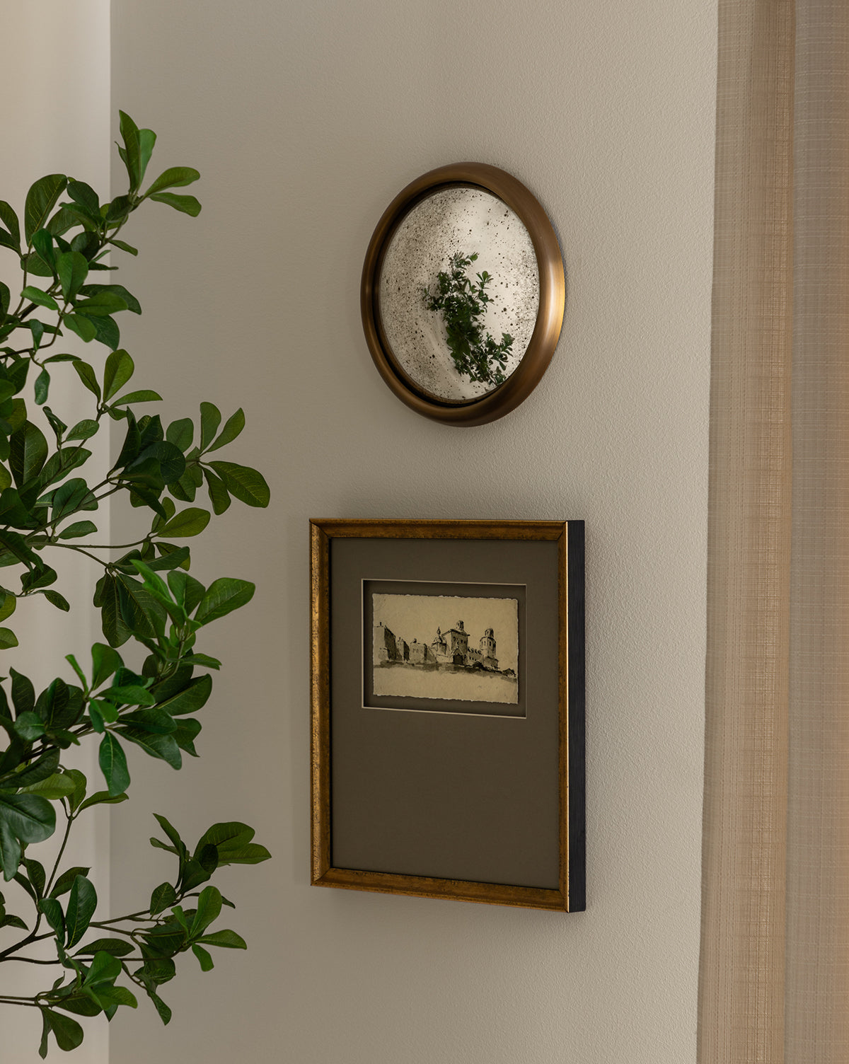 A leafy green plant stands near a beige wall adorned with the Juliette Convex Antiqued Mirror above a framed black-and-white architectural sketch, both in vintage brass frames, by McGee & Co.
