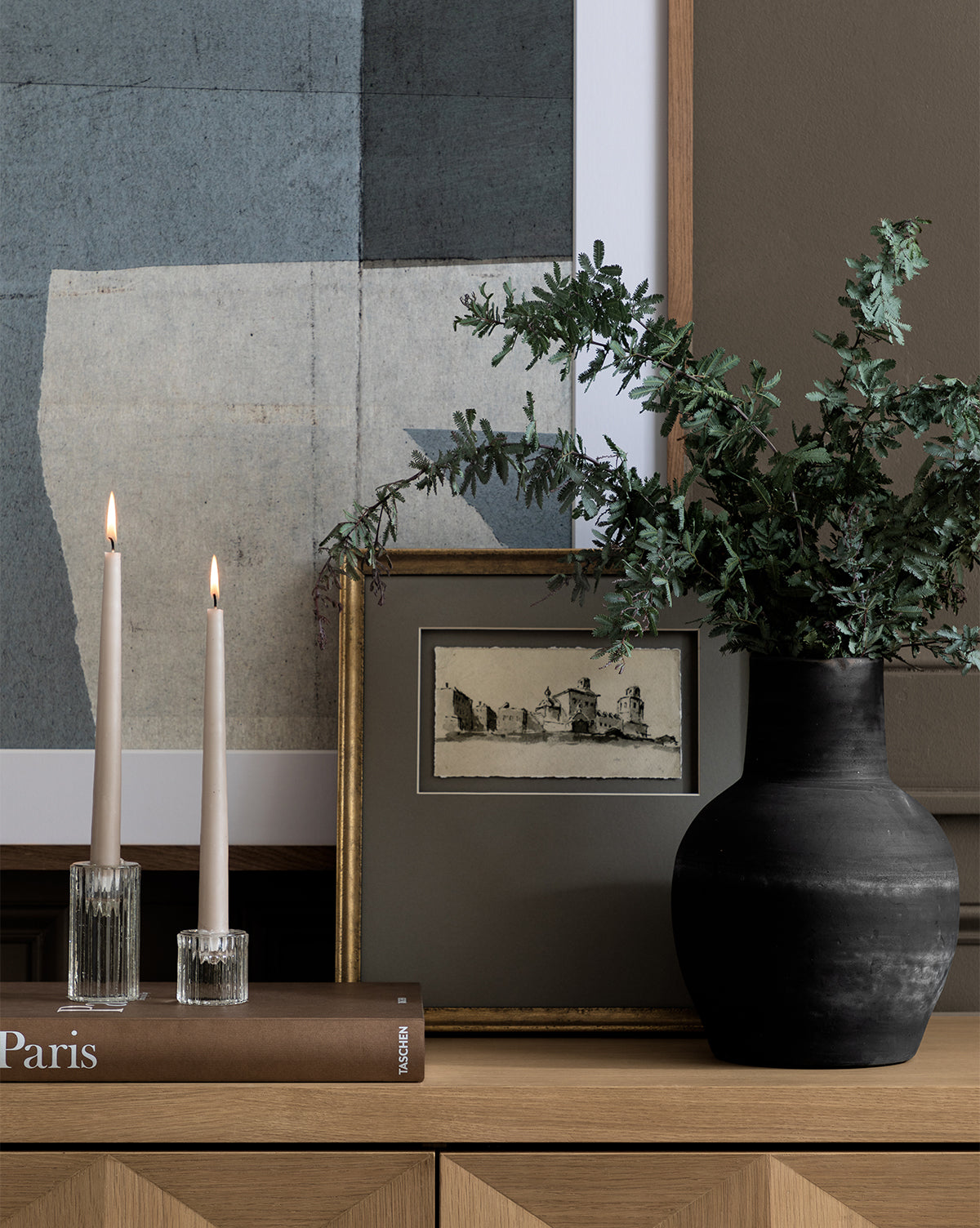 A wooden cabinet is adorned with the Auster Distressed Vase filled with greenery, two lit glass candles, a framed sketch, stacked books, and abstract artwork displayed on the wall behind, by McGee & Co.