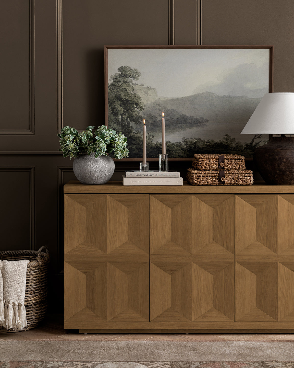 A wooden sideboard with geometric doors stands against a brown paneled wall, topped with candles, books, a potted plant, baskets, a ceramic lamp, and the Verde Landscape gallery-wrapped canvas. A woven basket sits on the floor beside it.