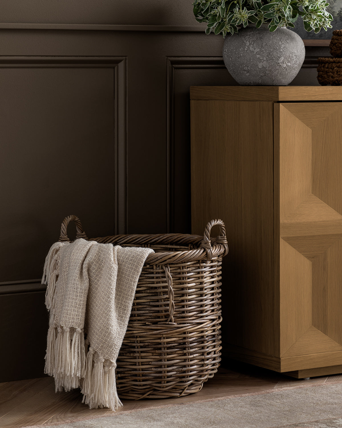 A Mckell Basket with handles holds a cream throw blanket by a wooden cabinet topped with a potted plant, all set against a dark brown paneled wall to showcase the natural beauty of the Mckell Basket.