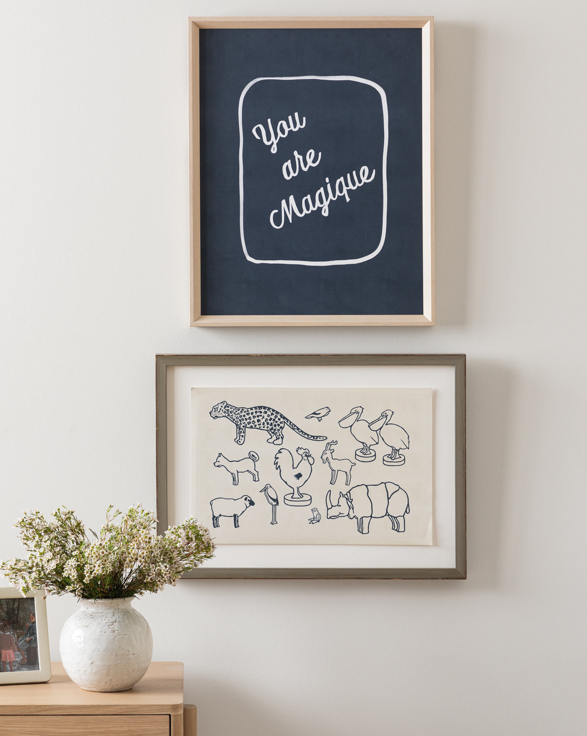 Two framed artworks hang above a light wood dresser with a white vase of flowers. The top frame features You are Magique, while the bottom displays simple animal line drawings similar to children’s art.