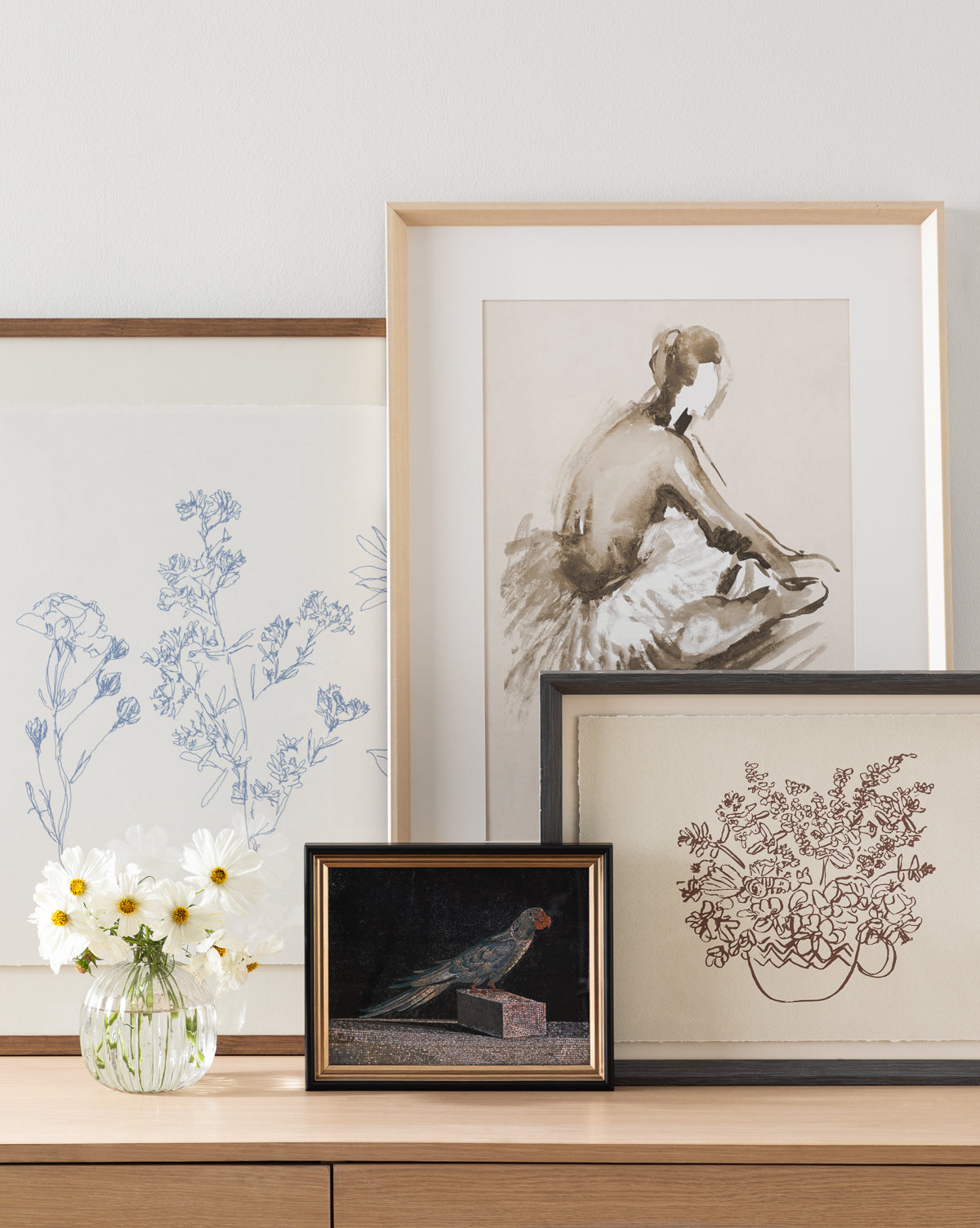 Blue Parrot, a framed art print, is displayed among drawings of flowers and a ballerina on a wooden surface. A glass vase with white daisies stands in front of the coastal-themed artwork.