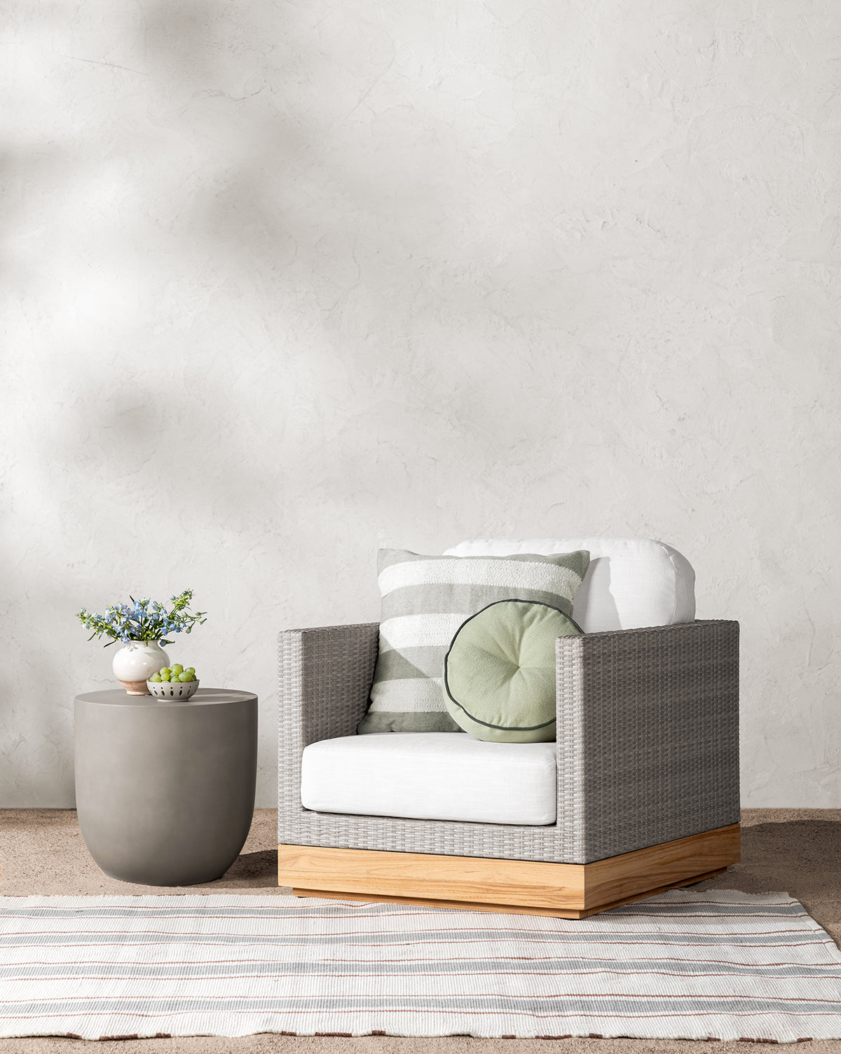 The Berdine Wicker Outdoor Chair with a white cushion and teak base sits beside a round gray side table with vases and flowers. Striped and round pillows accent the chair, all placed on a striped rug against a textured wall.