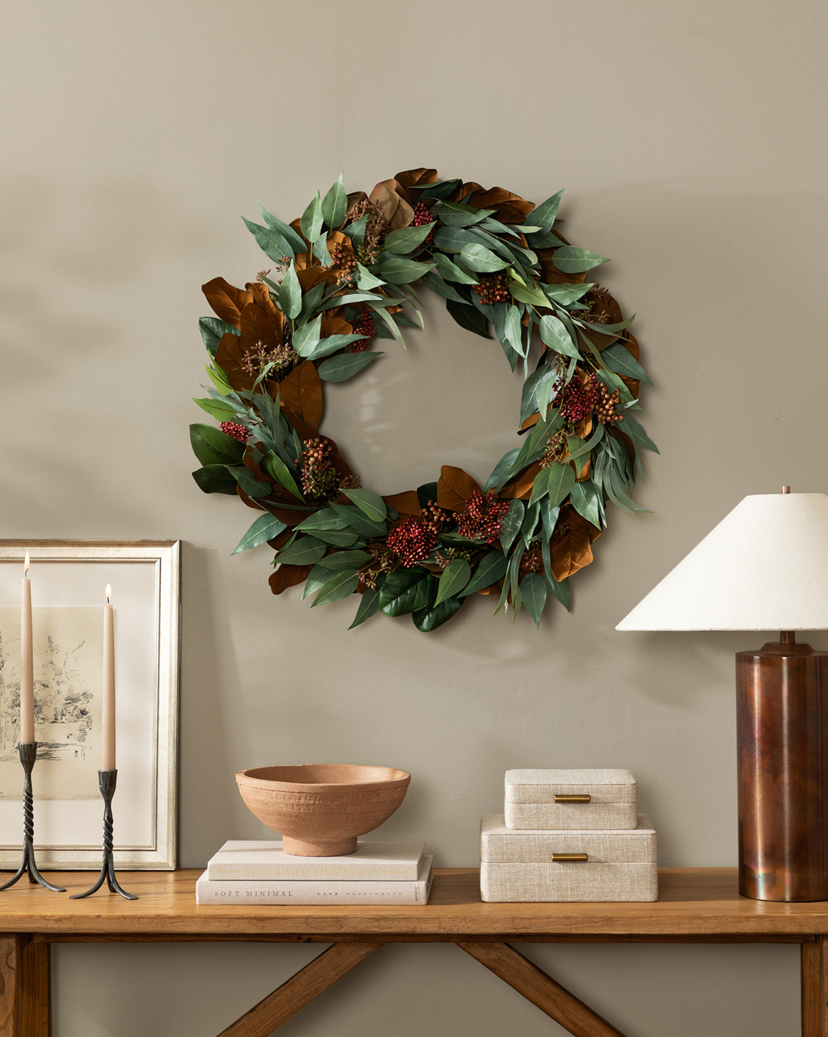 A 30 Faux Magnolia & Eucalyptus Wreath hangs on a beige wall above a wooden console table adorned with candles, books, a wooden bowl, stacked boxes, and a brown lamp with a white shade.