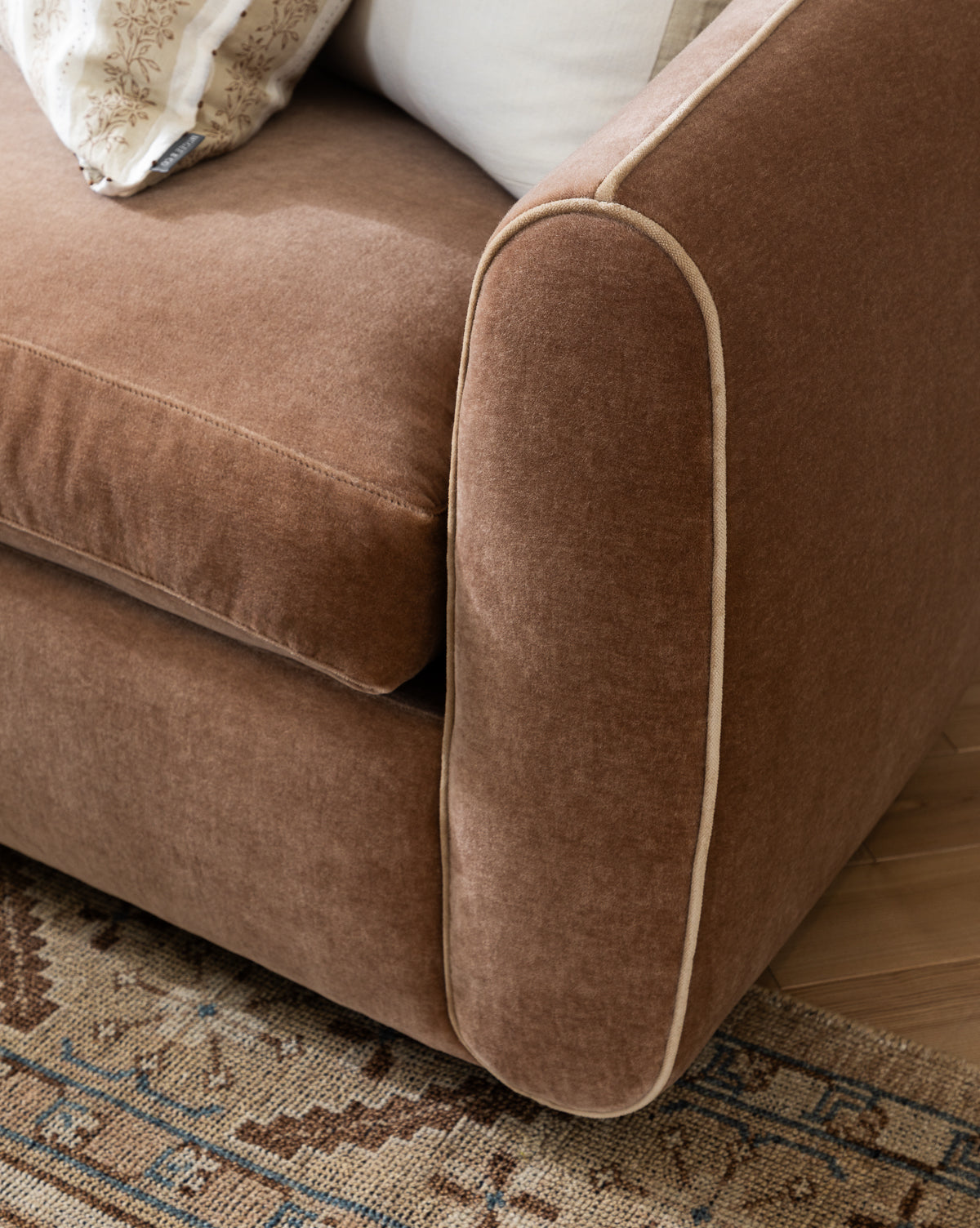 A close-up of the Wexler Sofa 90 in customizable designer fabric, shown with brown upholstery and light piping on the armrest, set on a patterned rug with cushions partially visible in the background, by McGee & Co.
