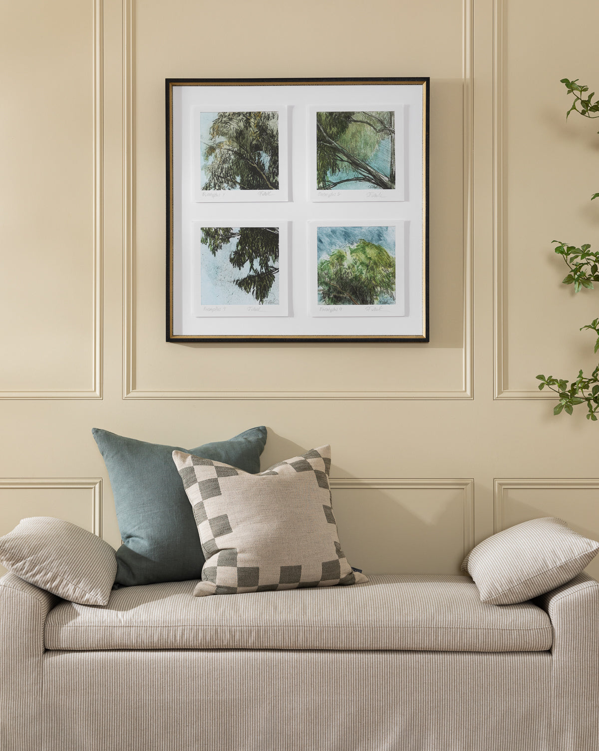 A beige sofa with patterned cushions sits against a cream paneled wall. Above, the Eucalyptus Windows giclee print hangs in a distressed black and gold frame, showing green trees and blue sky. A leafy plant appears on the right.