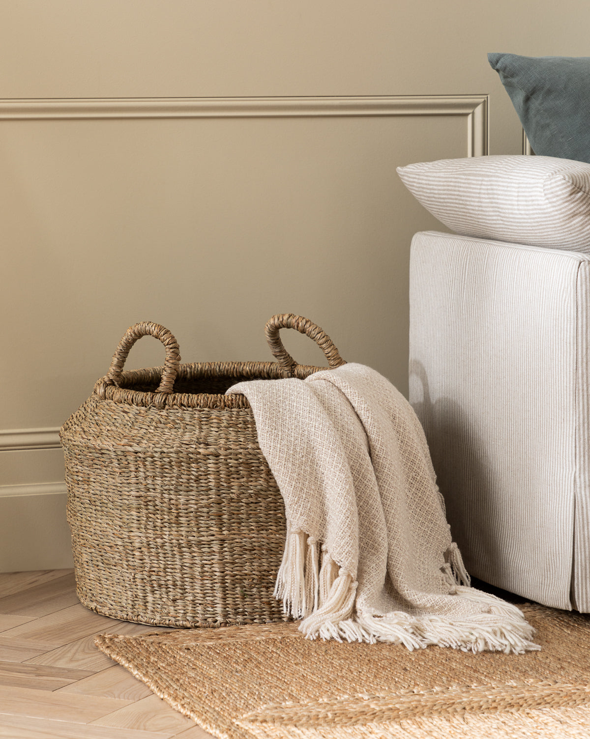 A Liesl Basket sits on a textured rug by a beige sofa, draped with a cream fringed blanket for a cozy accent in this neutral-toned living space, by McGee & Co.