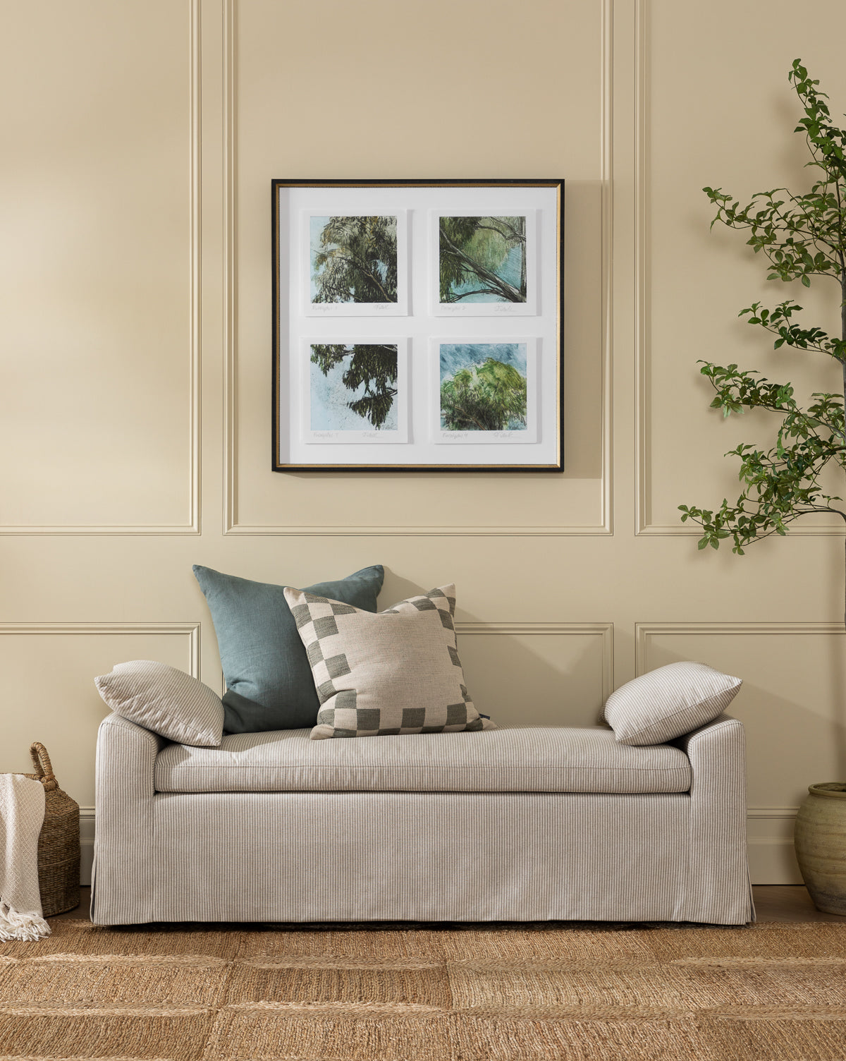 A neutral-toned living room with a light gray sofa, three cushions, the Scaletta Bench (Ready to Ship) in linen ticking stripe, a four-panel framed nature artwork, woven basket with blanket, green potted plant, and a textured rug by McGee & Co.