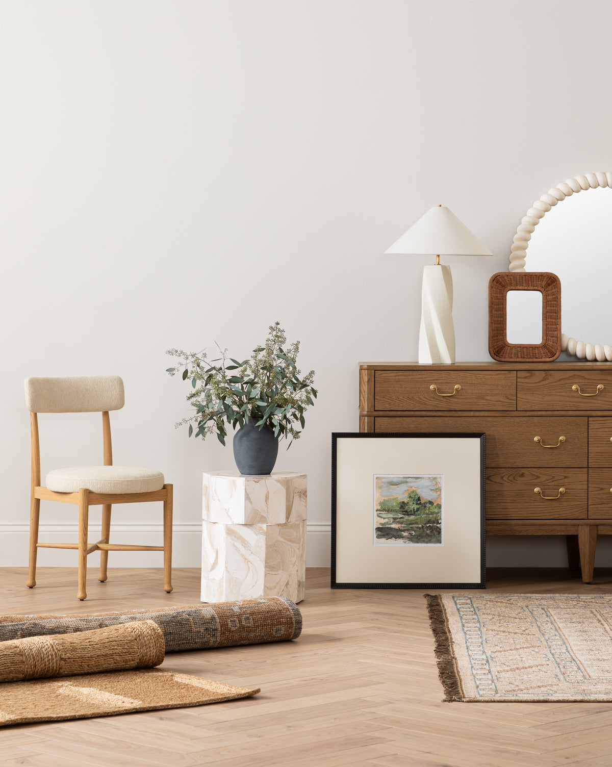 A minimal, modern room featuring a wooden chair, a vase with greenery on a patterned side table, a dresser with a lamp and mirror, and the Forest Meadow print in a black wood frame resting on the floor beside rolled-up woven rugs.