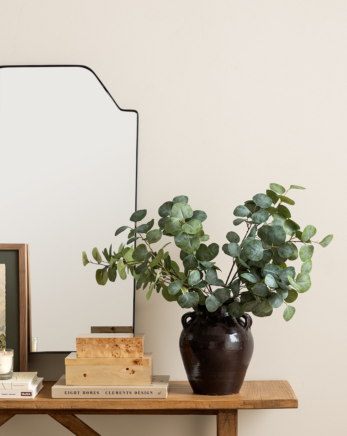 A dark vase with green eucalyptus branches sits on a wooden table next to stacked books, a Burl Wood Box, and a framed picture, with a mirror hanging on the beige wall behind for contemporary charm.