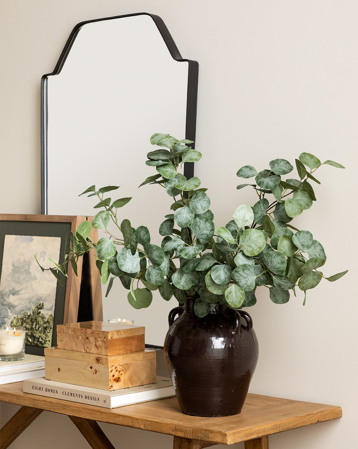A wooden table holds a black vase with a Faux Eucalyptus Branch, stacked books, a framed painting, a lit candle, and a decorative box, set before a wall-mounted mirror with a black-edged frame.