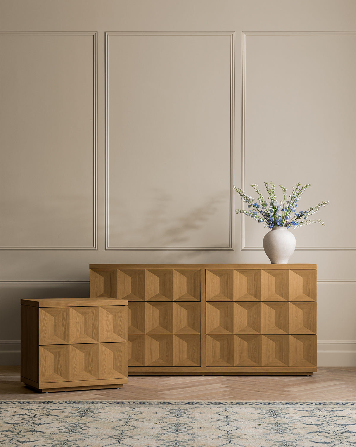 The Beacon Nightstand, featuring geometric patterns, stands against a beige paneled wall and offers chic bedroom storage; it is topped with a white vase holding blue and white flowers, while a patterned rug lies in the foreground, by McGee & Co.