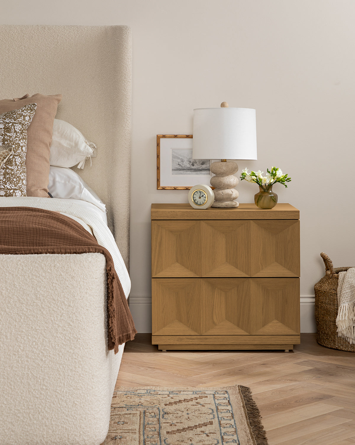 A cozy bedroom features a light upholstered bed with layered bedding beside the modern Beacon Nightstand, which holds a lamp, clock, framed photo, and small vase of flowers. A woven basket on the floor adds extra storage by McGee & Co.