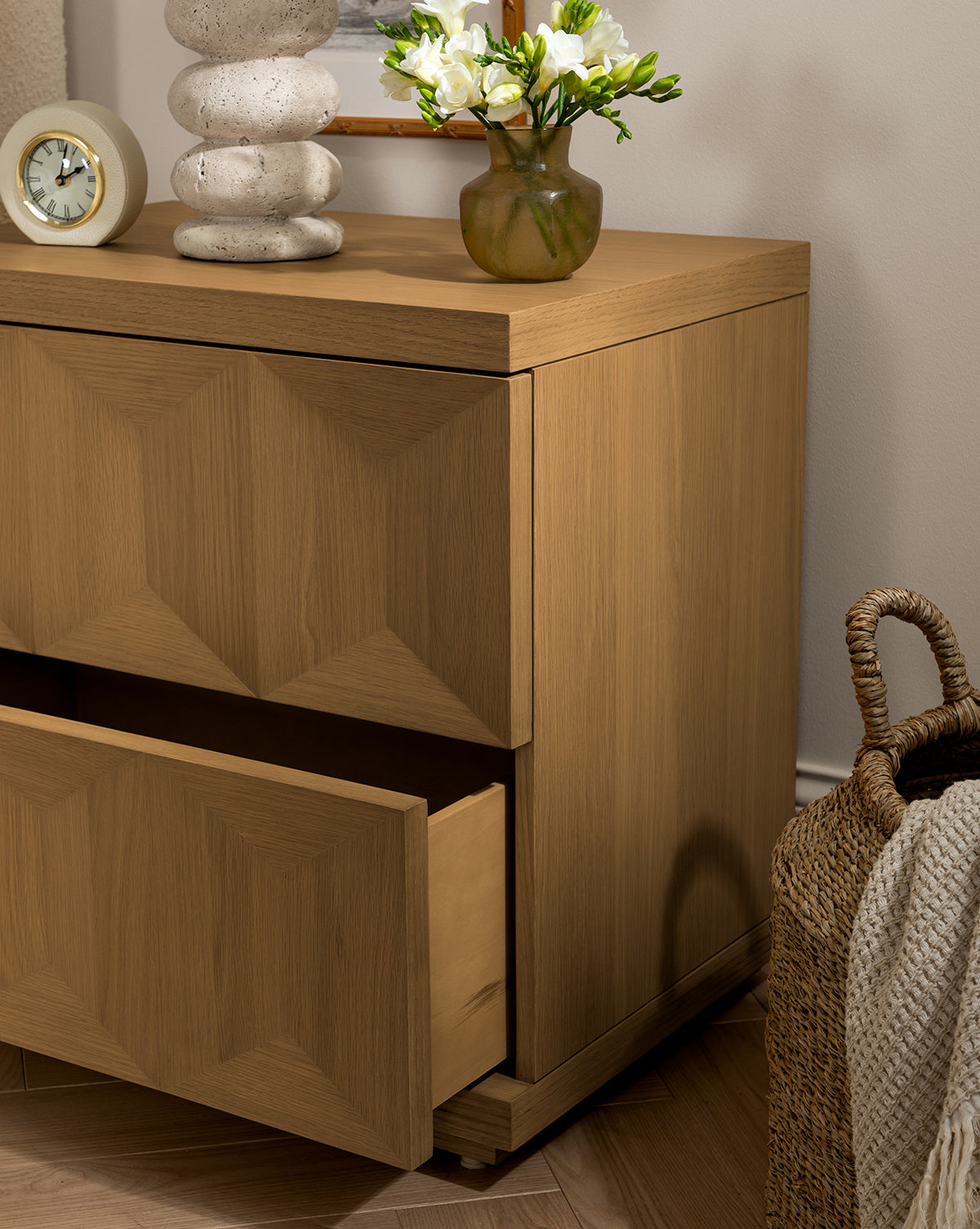 The Beacon Nightstand stands against the wall, providing bedroom storage. On top rest a small clock, a stone-like lamp, and a vase with white flowers, while a woven basket sits on the floor beside this modern piece by McGee & Co.