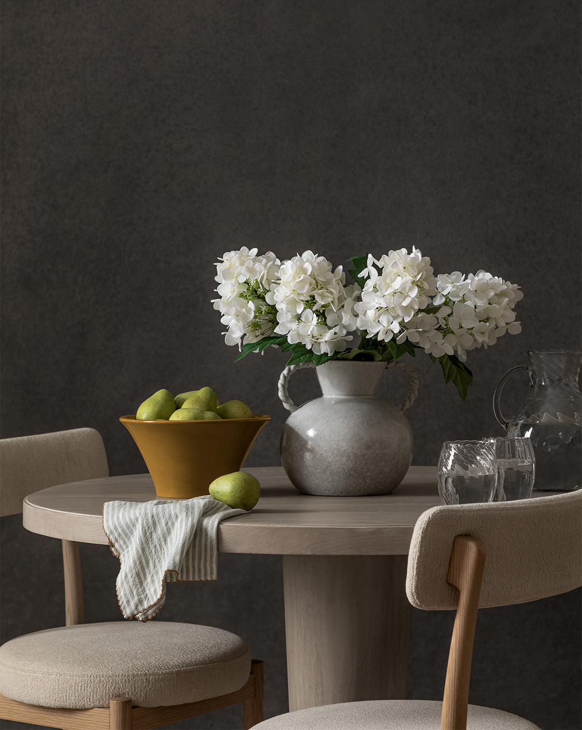 A round wooden table with two beige chairs displays a vase of white flowers, the Brennan Serving Bowl filled with green pears, a striped cloth, two empty glasses, and a glass pitcher as elegant kitchen decor against a dark background, by McGee & Co.