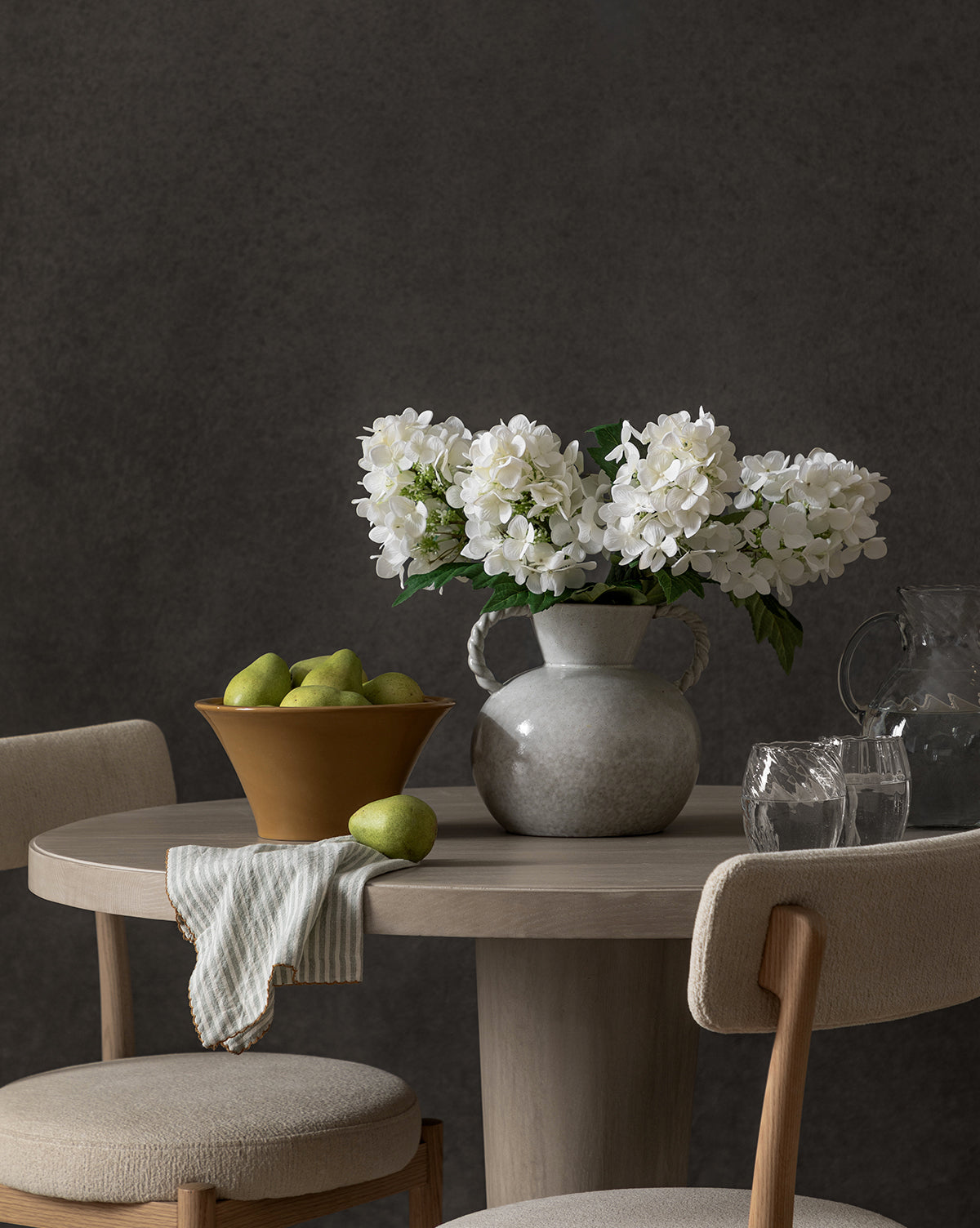 A round wooden table displays a Faux Hydrangea Stem in a vase, a brown bowl of green pears, striped cloth, three clear glasses, and a glass pitcher. Two upholstered chairs complete the scene against a dark gray background.