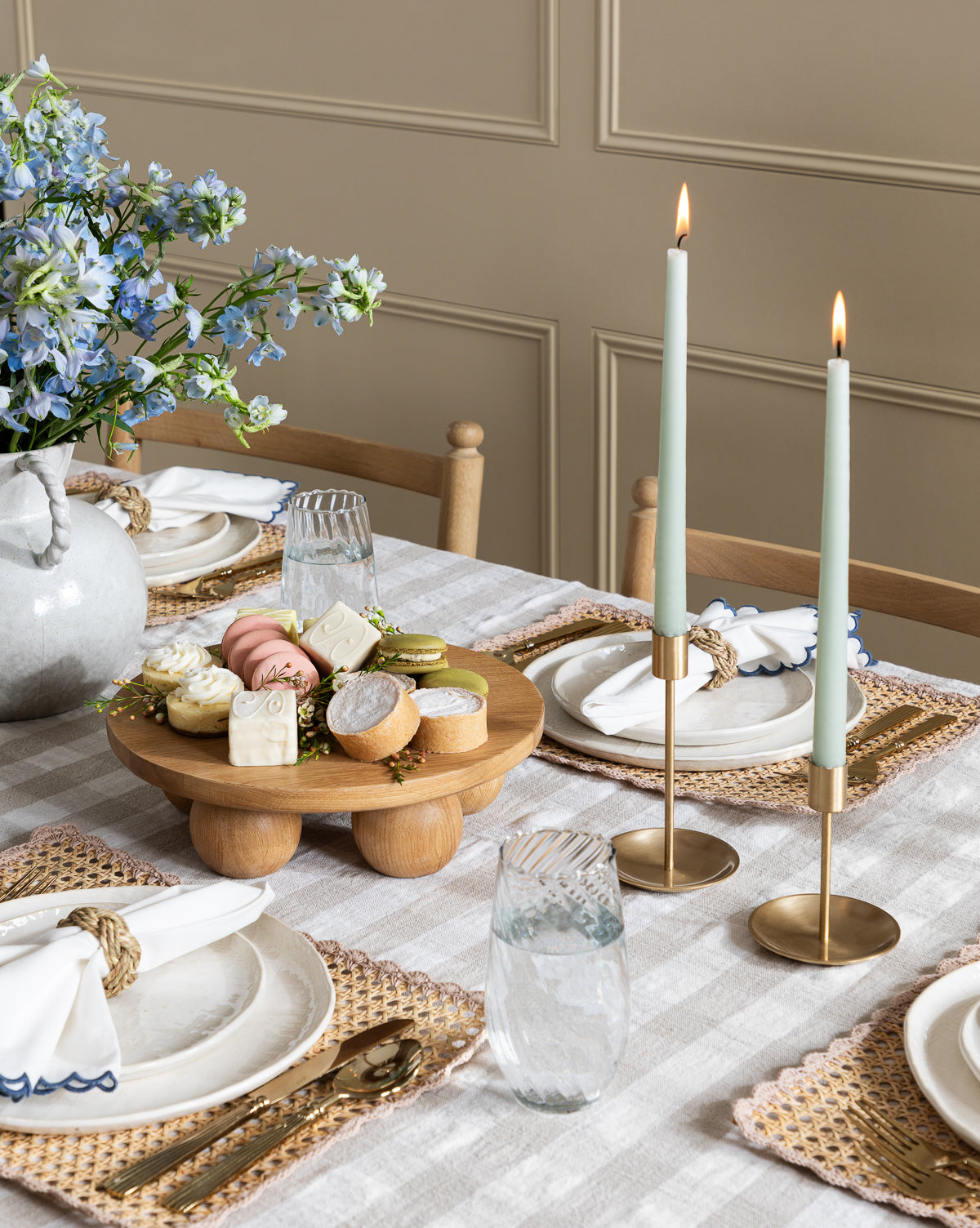 A dining table set with woven placemats, white plates, glasses, rope-napkin rings, a vase of blue flowers, two lit candles in Brass Taper Holders, and a wooden tray with assorted desserts and macarons.