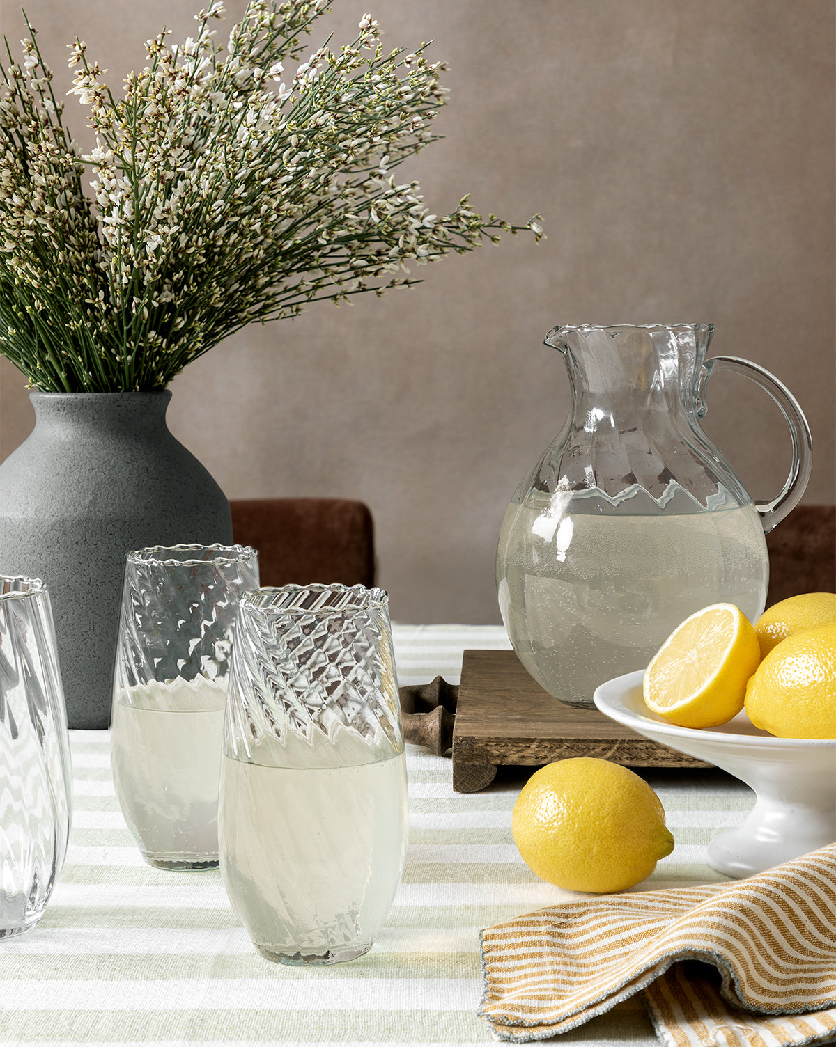 An Adderley Pitcher and three scalloped rim glasses of lemonade rest on a striped tablecloth, accompanied by a plate of lemons, a yellow-striped napkin, and a vase of white flowers, by McGee & Co.