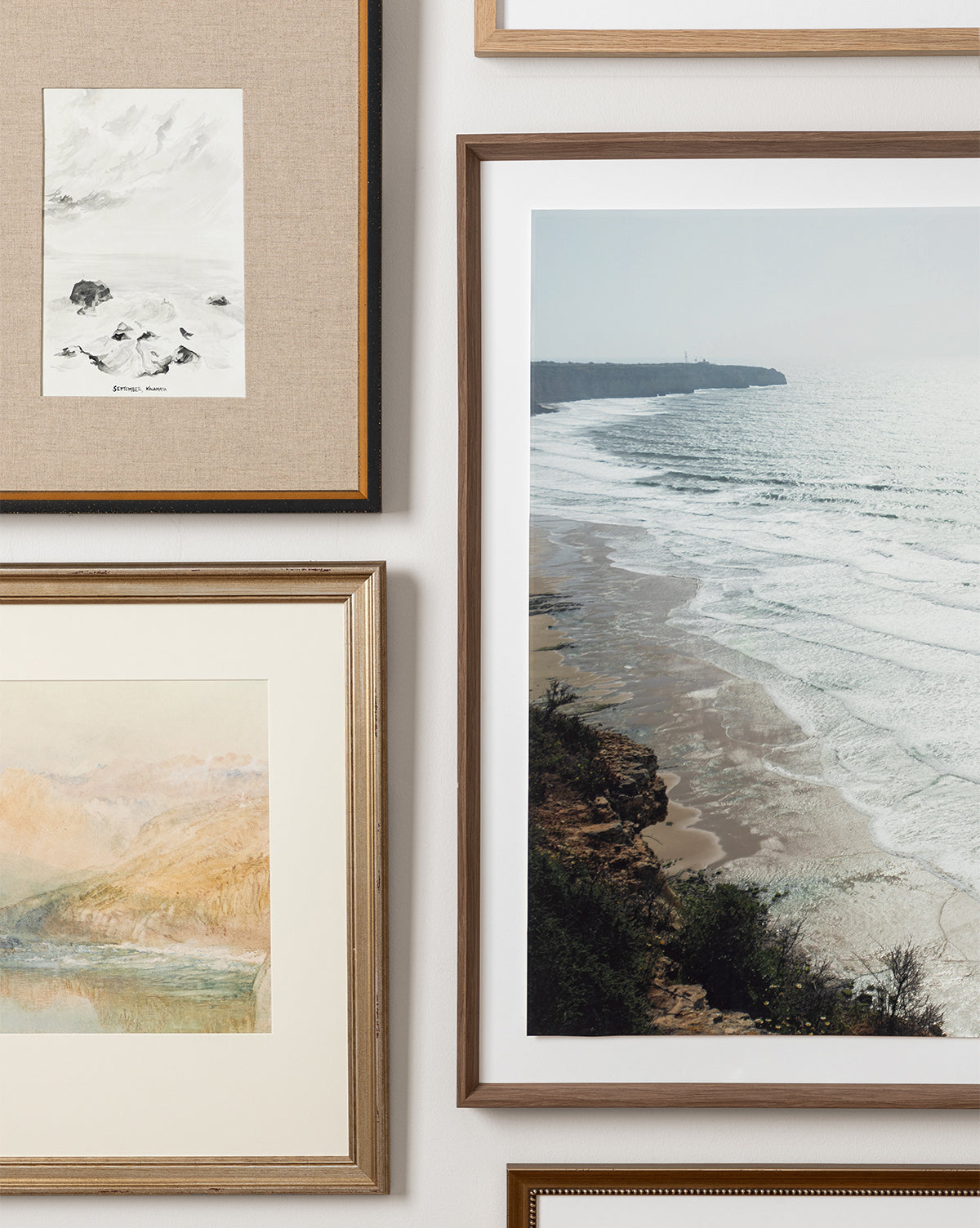 A close-up of a gallery wall with framed art, featuring Sunset Hills, a large giclee coastline print with waves, alongside a watercolor landscape and a black-and-white sketch on neutral backgrounds.