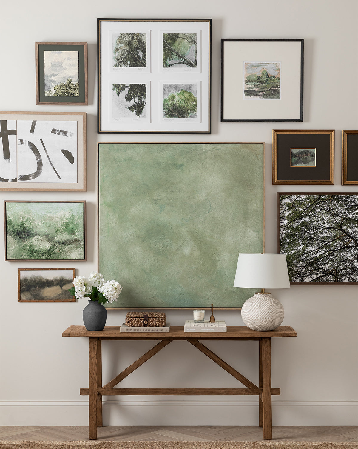 A wooden console table holds a white lamp, books, and flowers beneath framed art, including landscapes, abstracts, botanicals, and the striking Treetops giclee print in a natural wood frame.