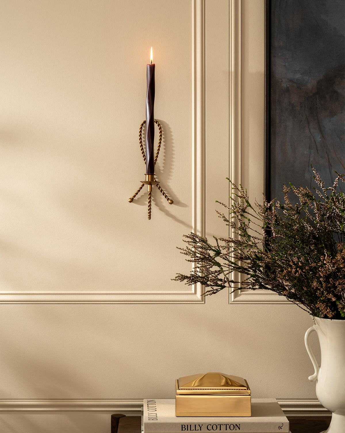 A lit candle in the Hearst Taper Holder is mounted on a cream paneled wall. Below, a stack of books and a gold box rest on a table beside a white vase with dark greenery, by McGee & Co.