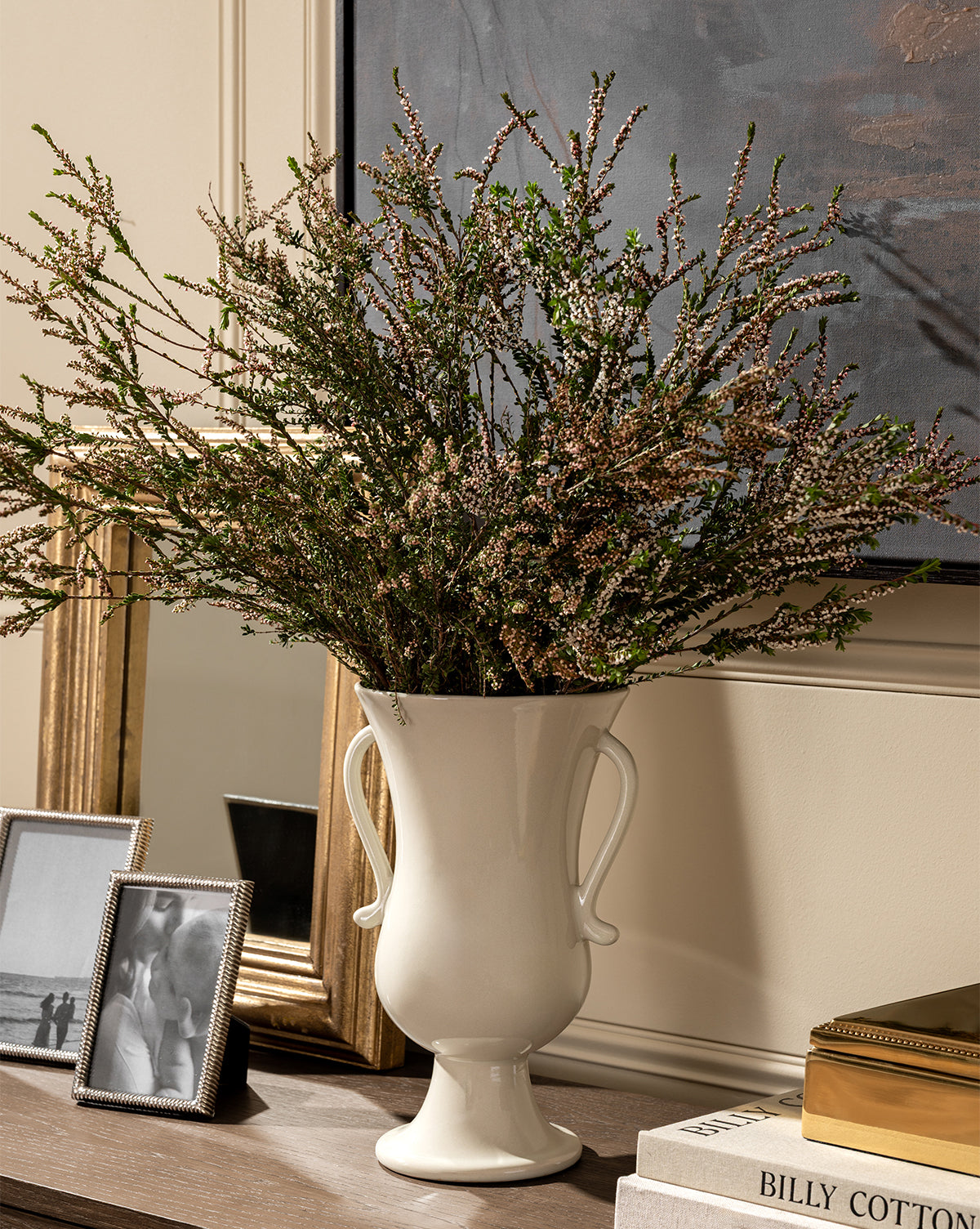 The Delphine Footed Vase, a tall and elegant white piece with handles, displays greenery on a wooden surface beside framed photos, books, and a gold-bordered mirror, with part of a painting visible in the background, by McGee & Co.