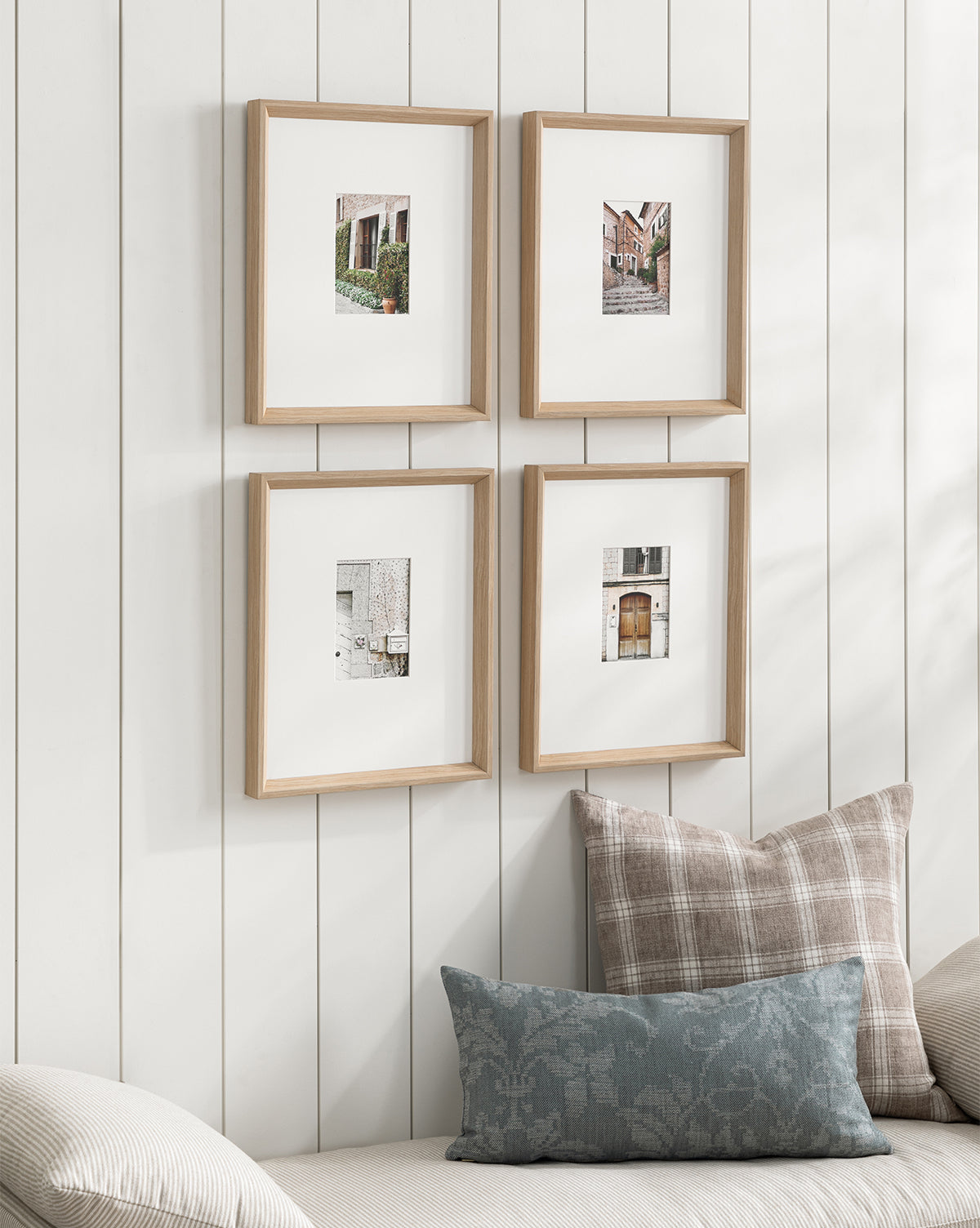 Four Valldemossa natural woodgrain frames with white mats and small photos are arranged in a grid on a white paneled wall above a sofa with neutral plaid and patterned pillows, creating a bright and cozy setting.