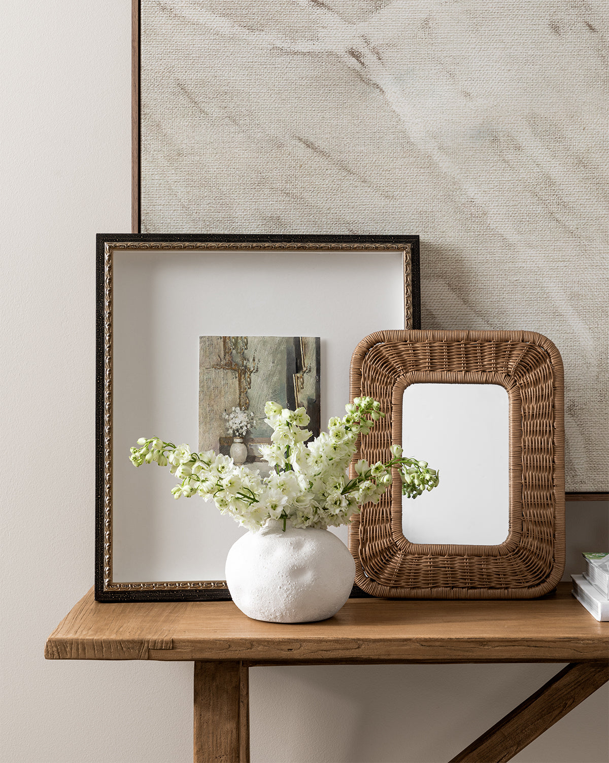 Golden Still Life features a wooden table styled with a white vase of flowers, a framed floral giclee print, an empty frame, and a woven rattan mirror for an elegant display against a neutral textured wall.