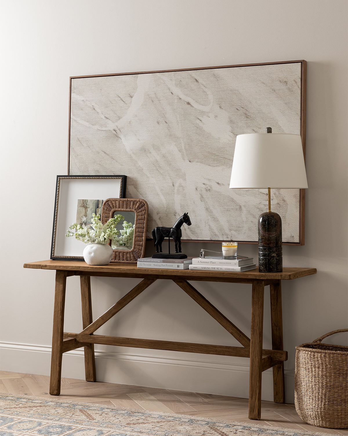 A wooden console table with a lamp, books, framed art, white flower vase, black horse figurine, and candle sits against a neutral wall featuring The Oath 2 by Shatha Al Dafai as large wall art above.