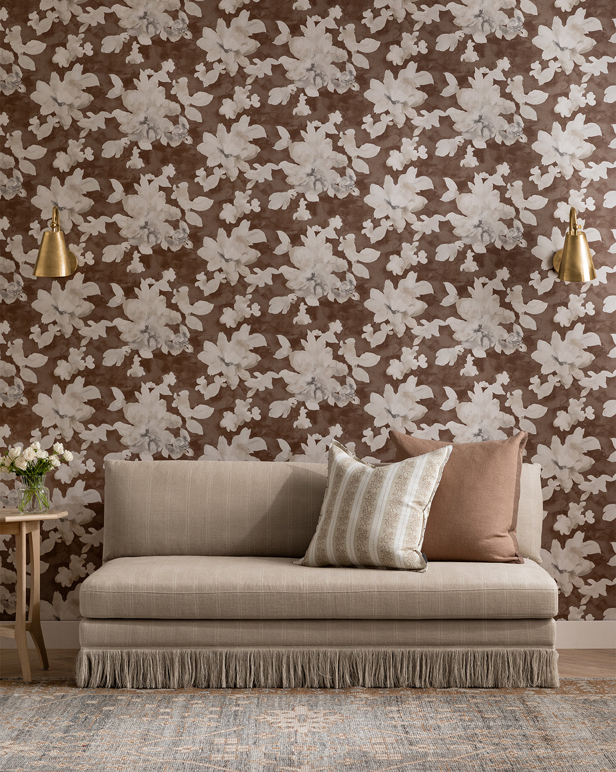 The Philippa Settee (Ready to Ship), a beige modern upholstered piece with fringe trim, sits against brown floral wallpaper. Two pillows, gold wall sconces, and a side table with flowers complete the cozy, elegant setting, by McGee & Co.