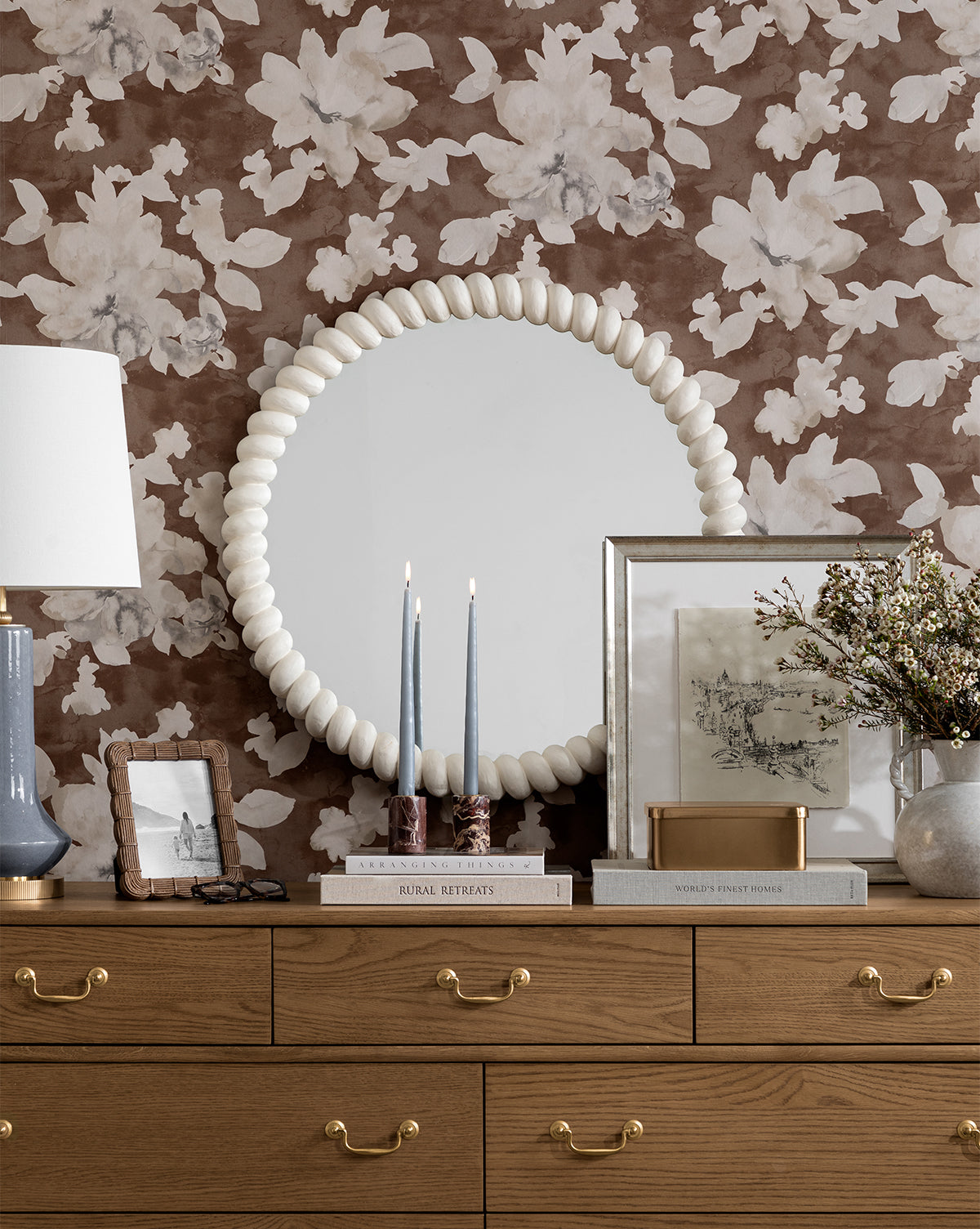 A wooden dresser with gold handles holds books, framed photos, candles, a vase of flowers, and a lamp. Above it, a round beaded-frame mirror hangs on brown floral wallpaper beside Etched City, an artwork depicting a city scene.