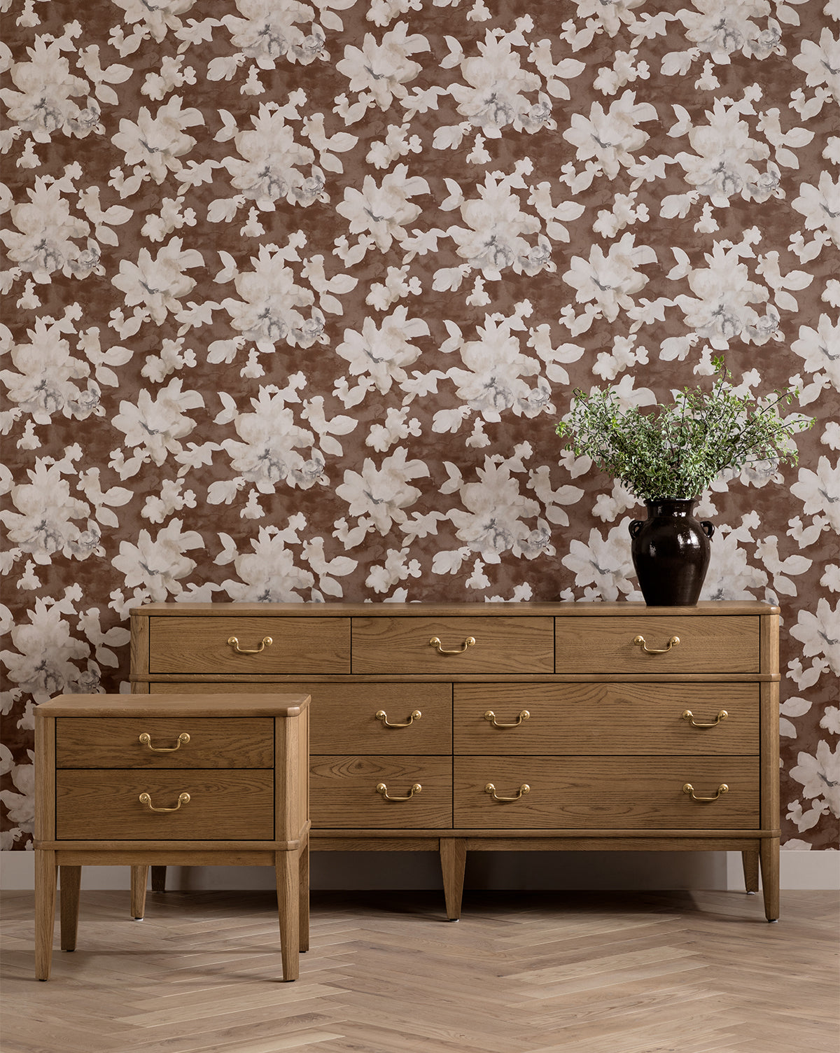 An Alessandra Nightstand with brass handles and a wooden dresser stand on a herringbone floor. A vase with green leafy branches and brown floral wallpaper complete the cozy scene, by McGee & Co.