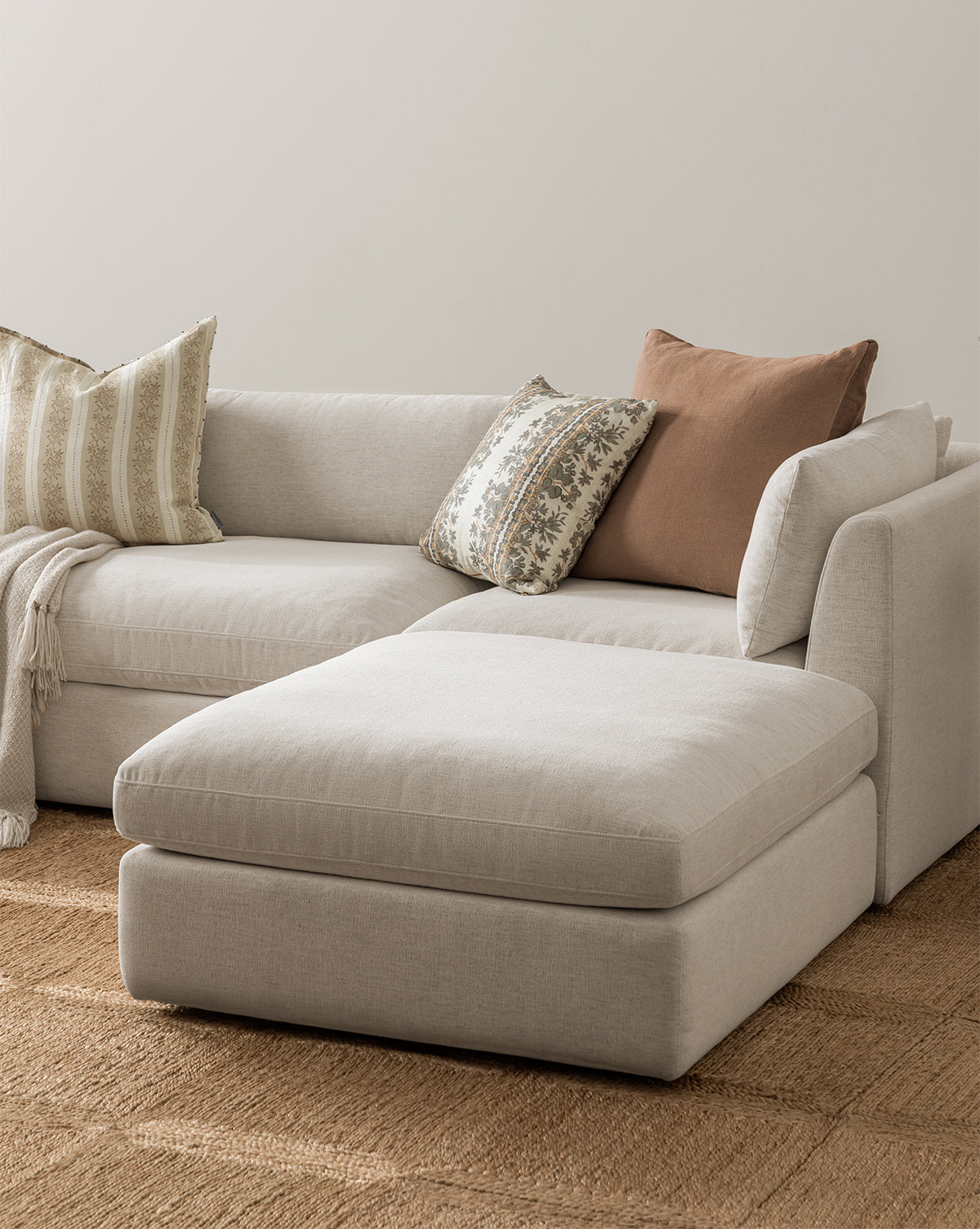 A light beige sectional sofa with an extended chaise sits on a woven rug beside the Vivienne Modular Ottoman. Two patterned throw pillows and a solid brown pillow are on the sofa, with a soft blanket draped over the armrest, by McGee & Co.