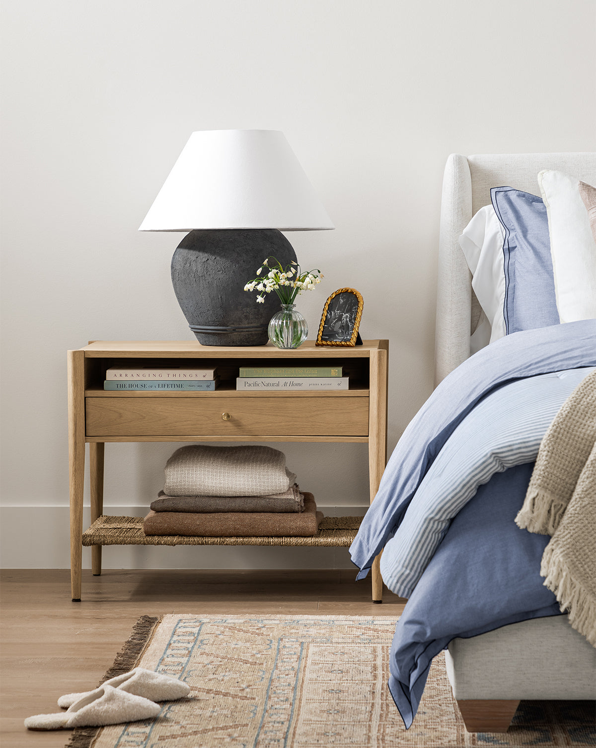 The Dana White Oak Nightstand with brass hardware holds a white lampshade, books, a small vase of flowers, and a framed photo beside a bed with blue and white bedding, by McGee & Co. Folded blankets are neatly stored on its woven seagrass shelf below.