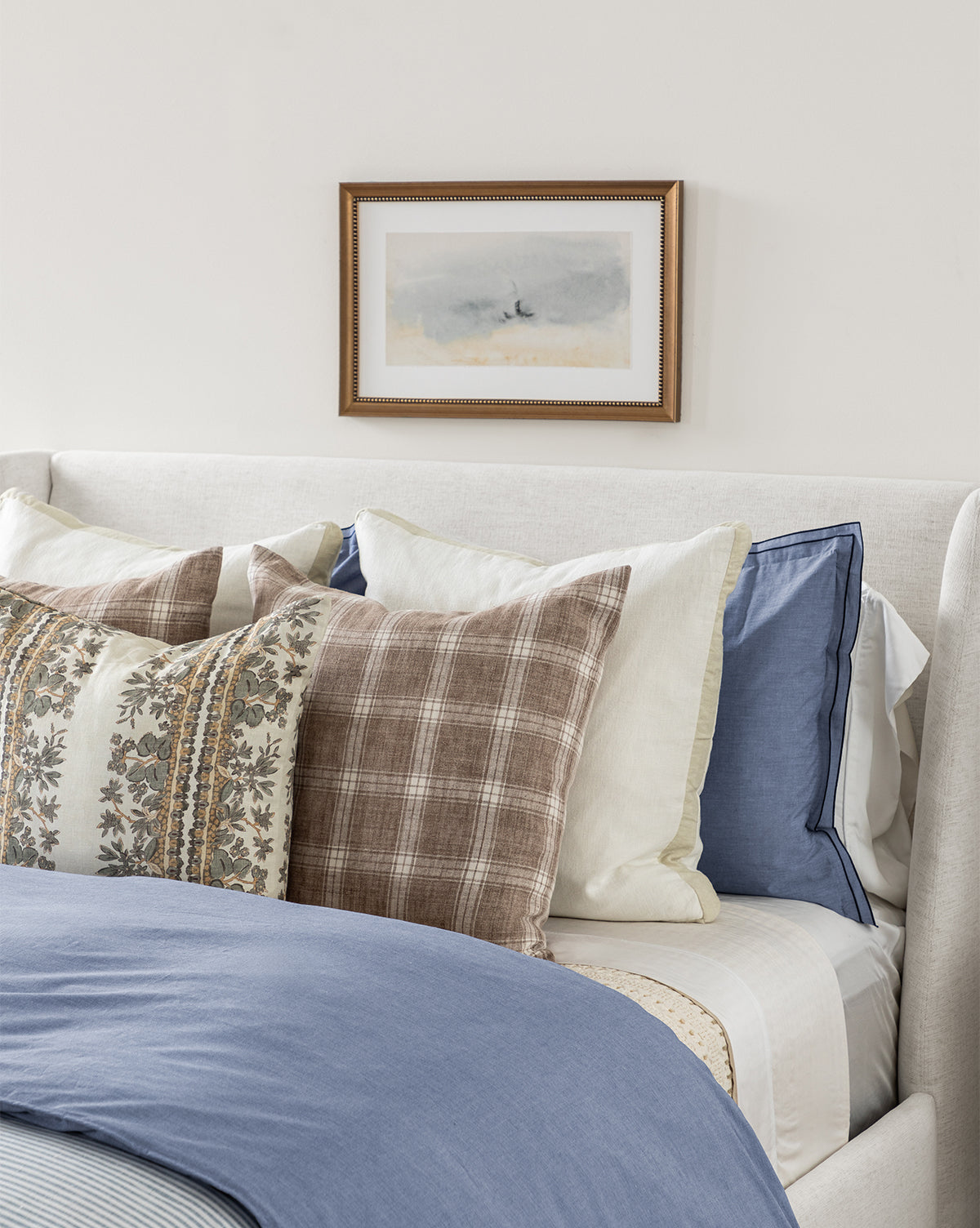 A neatly made bed with layered pillows in blue, plaid, floral, and cream patterns is topped with a light blue comforter. Above the bed hangs the Sky Studies 4 giclee print in an antique silver wood frame on a white wall.