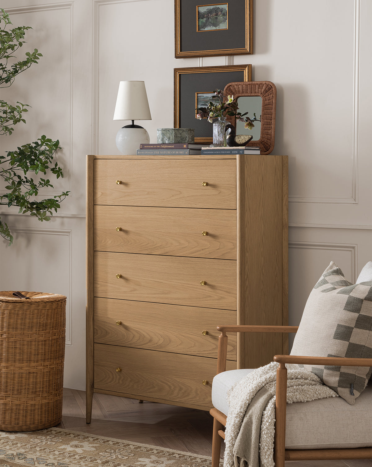 The Dana Tallboy Dresser with brass hardware stands against a white paneled wall, topped with a lamp, books, plant, and mirror. Framed art hangs above, while a chair and woven basket sit nearby with a leafy plant on the left by McGee & Co.