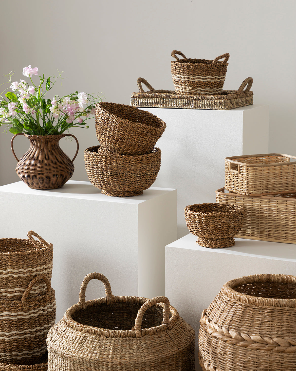 A group of woven baskets in assorted shapes and sizes are displayed on white pedestals beside the Antonia Woven Vase, which holds pink and white flowers against a neutral wall.