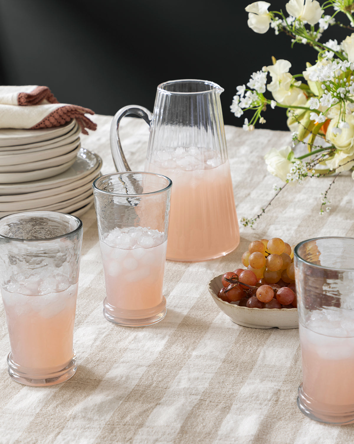 The Adonis Ribbed Glass Pitcher with three pink lemonades on ice sits on a striped tablecloth beside grapes, stacked plates with napkins, and a vase of white flowers for a fresh kitchen look, by McGee & Co.